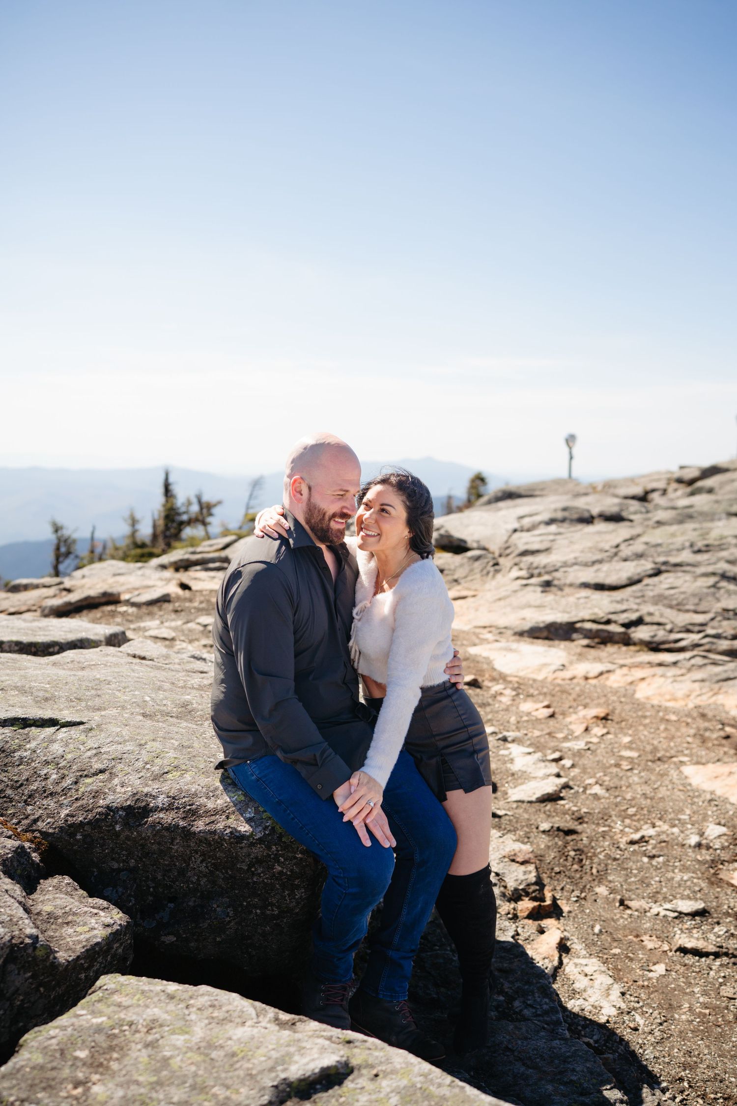 Couple sharing a romantic moment on rocky mountain summit, woman smiling as man embraces her on a clear sunny day.