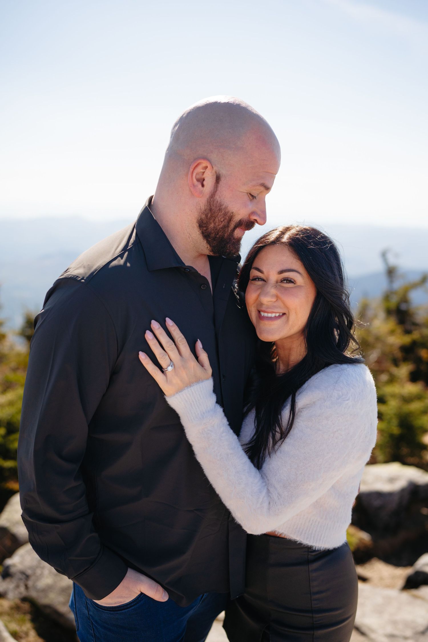 Couple embracing on a mountain summit, woman smiling with engagement ring visible, scenic sky background.