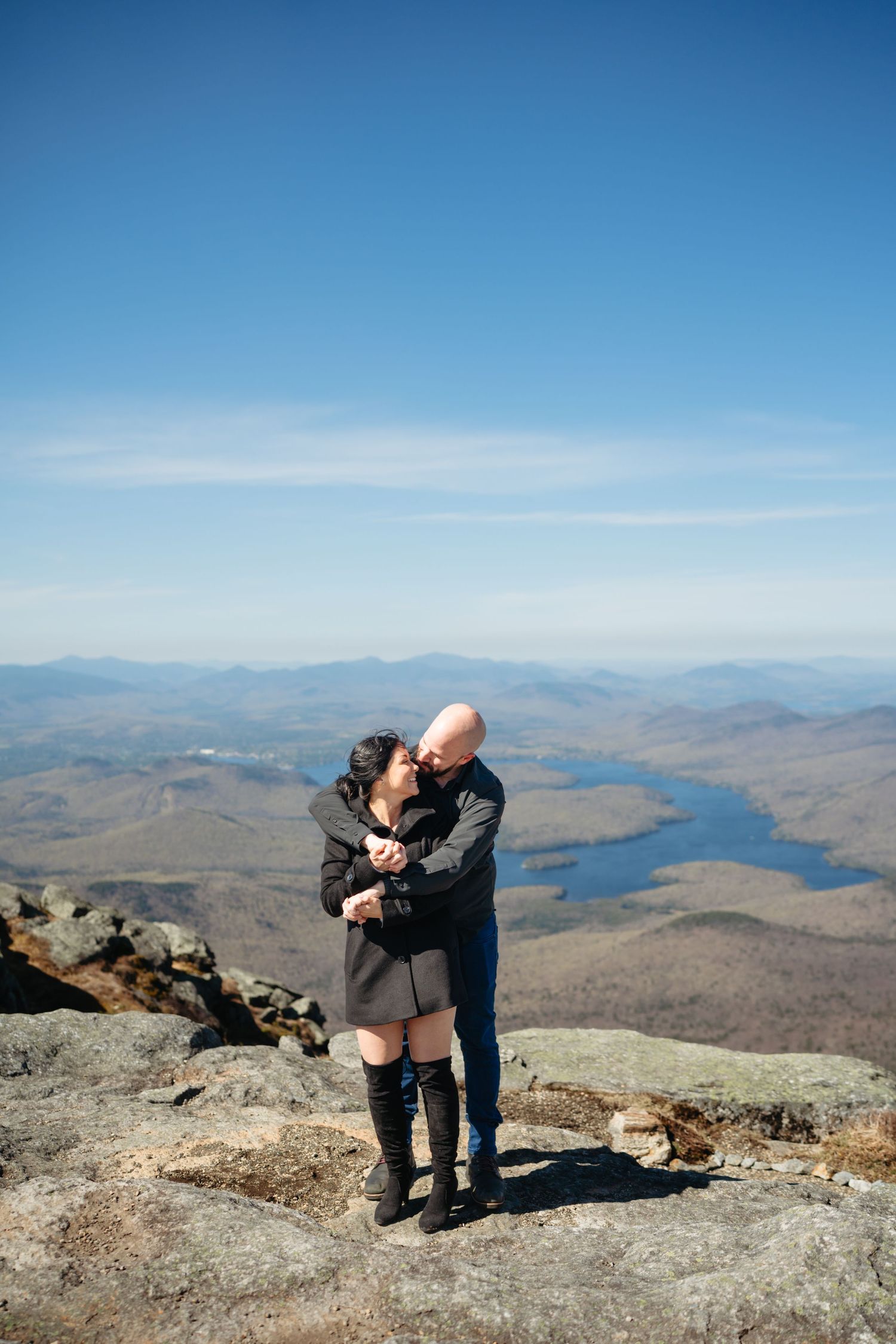 Couple embracing on a mountain summit with stunning aerial views of lakes and autumn foliage below on a clear day.