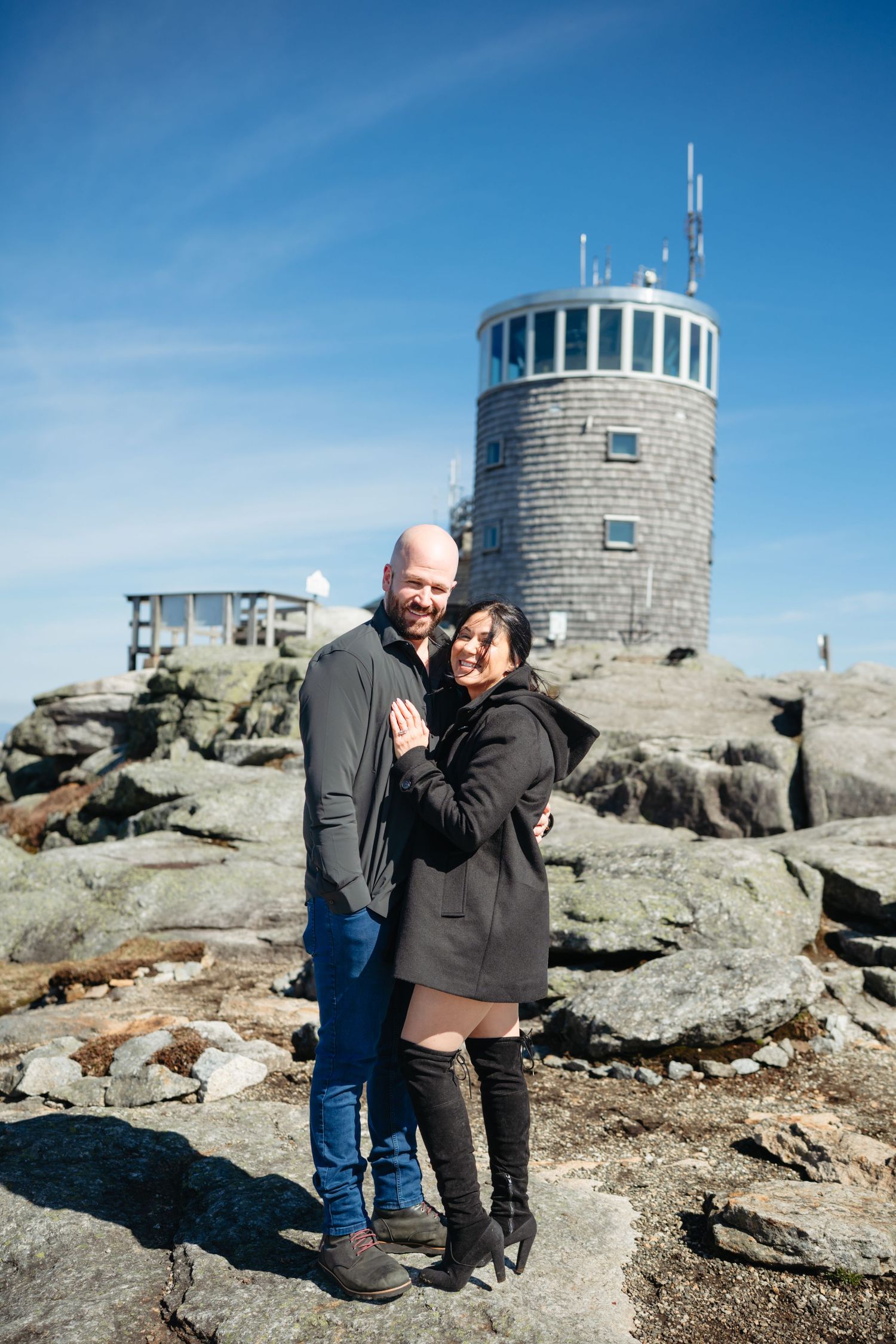 Couple embracing on rocky mountain summit with historic stone observation tower and blue sky backdrop.