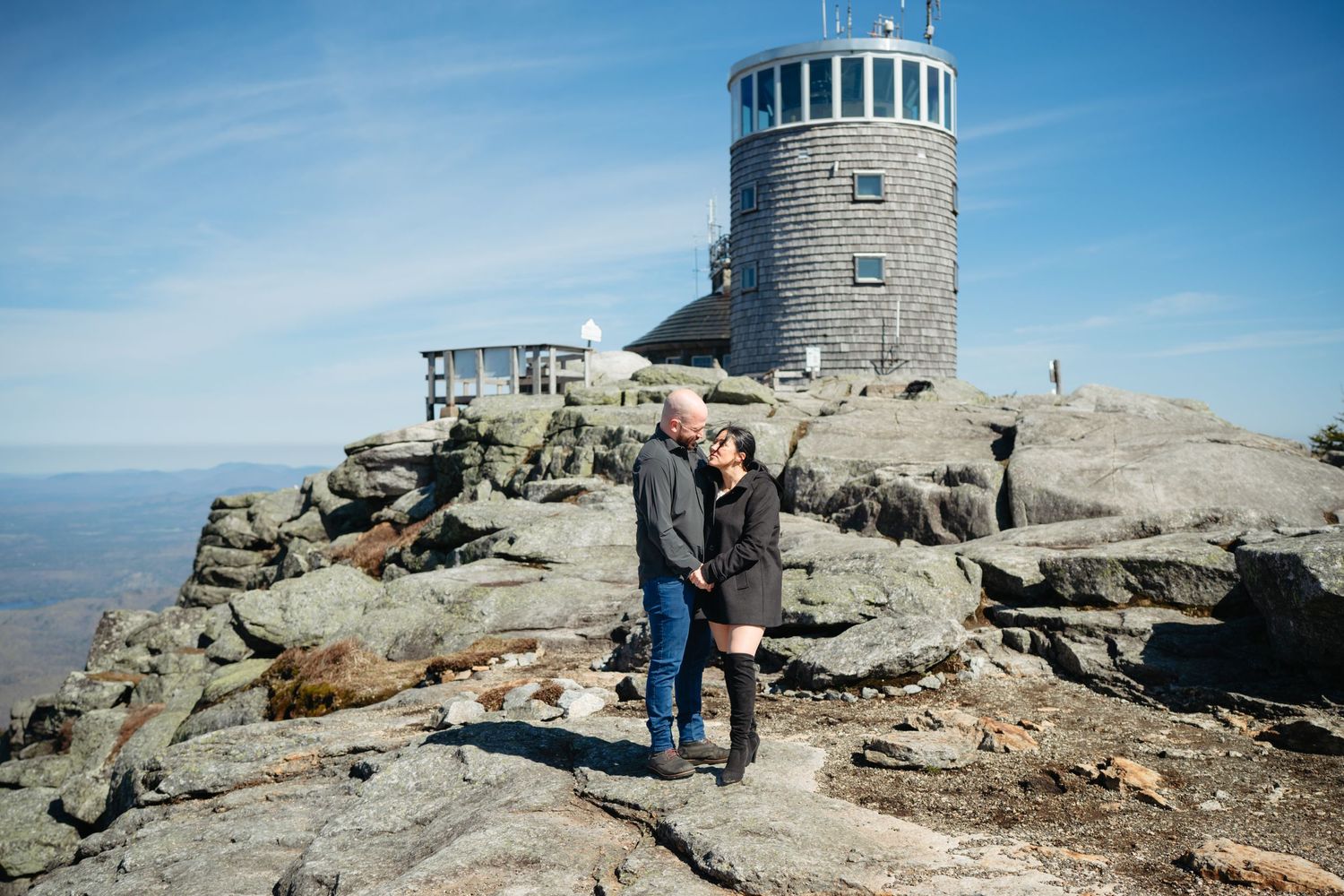 Couple embracing on rocky mountain summit with historic stone observation tower and blue sky backdrop.
