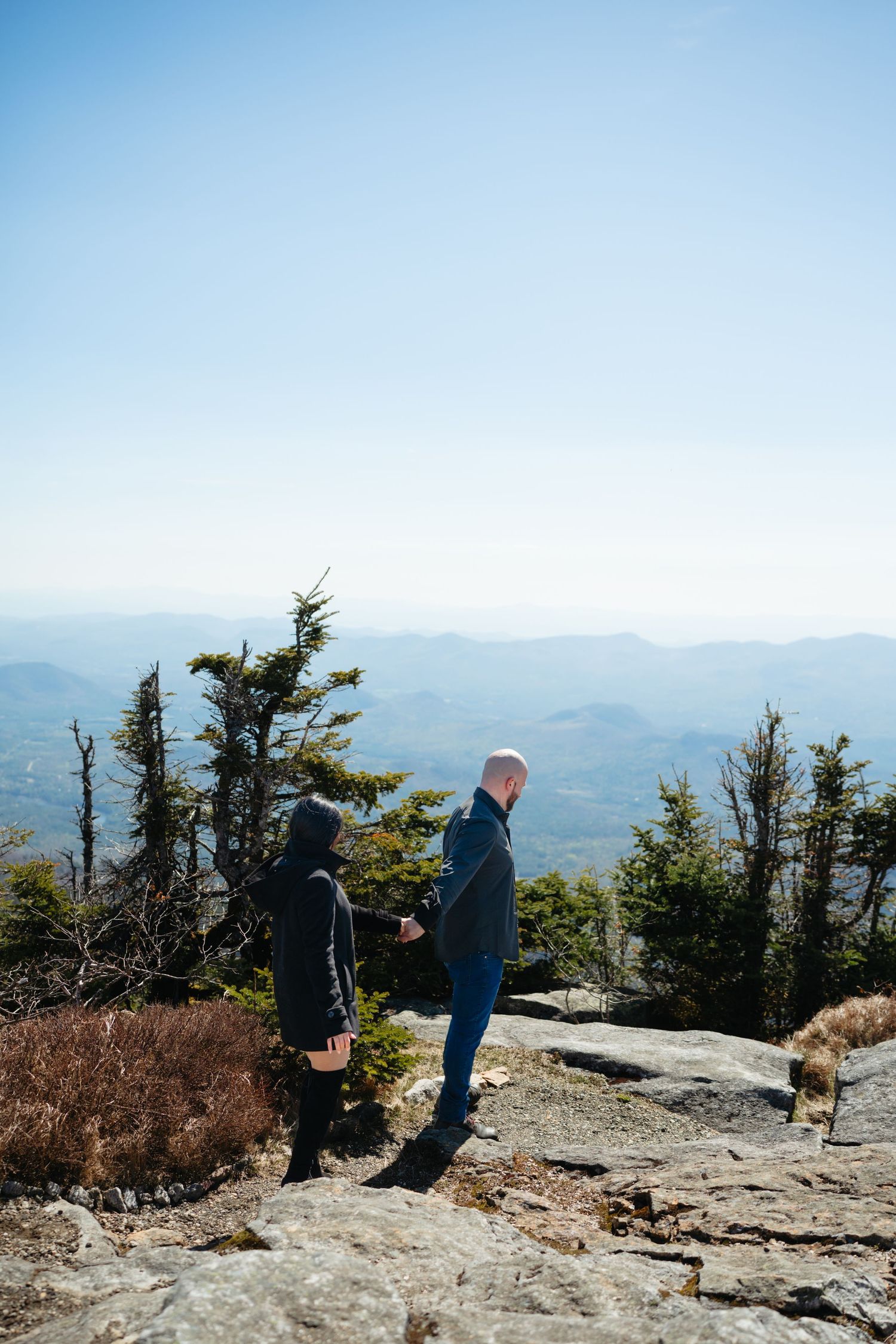 Couple holding hands on a rocky mountain overlook surrounded by evergreen trees and vast mountain views.