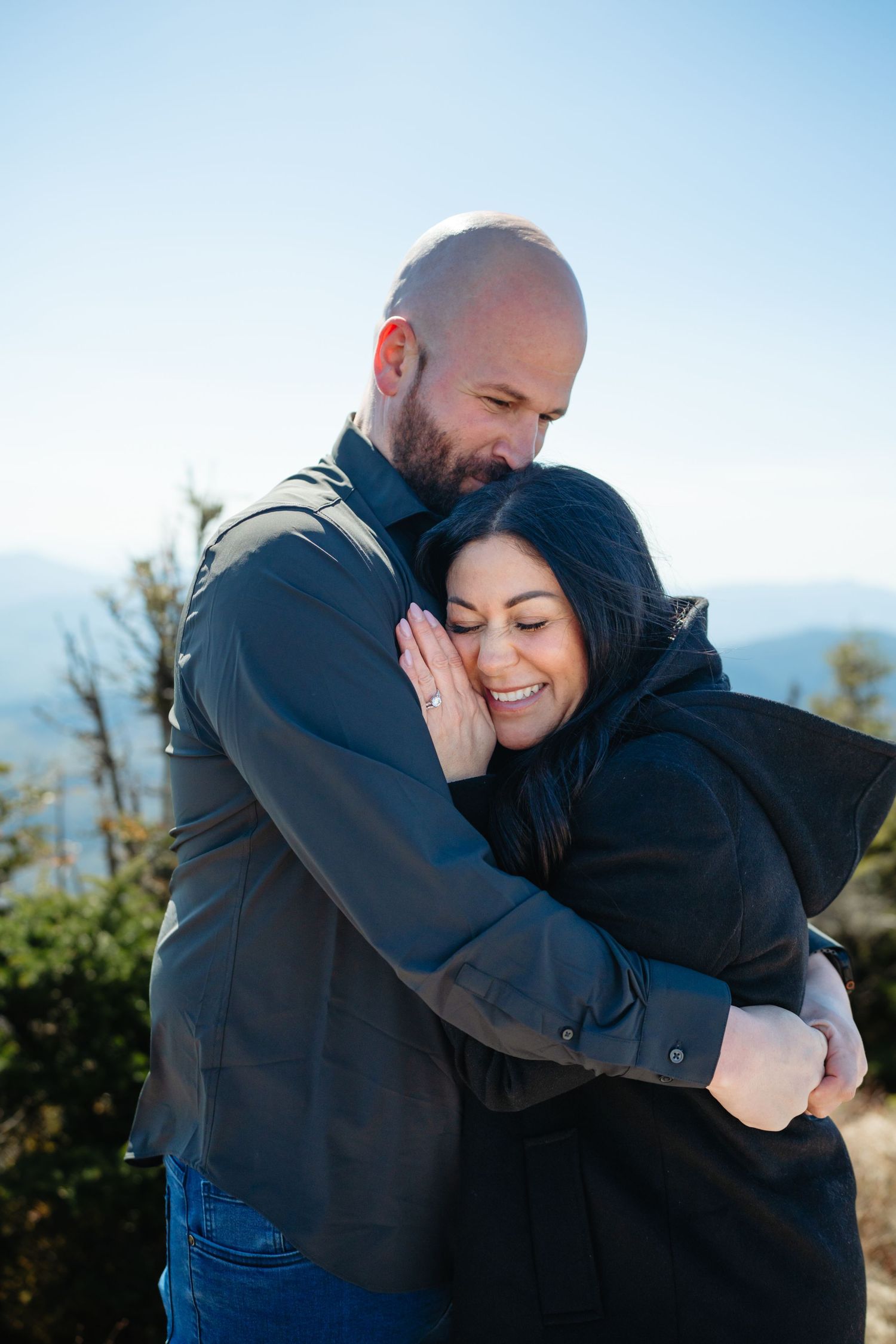 A bald man with a beard embraces a smiling dark-haired woman outdoors with mountains in the background.