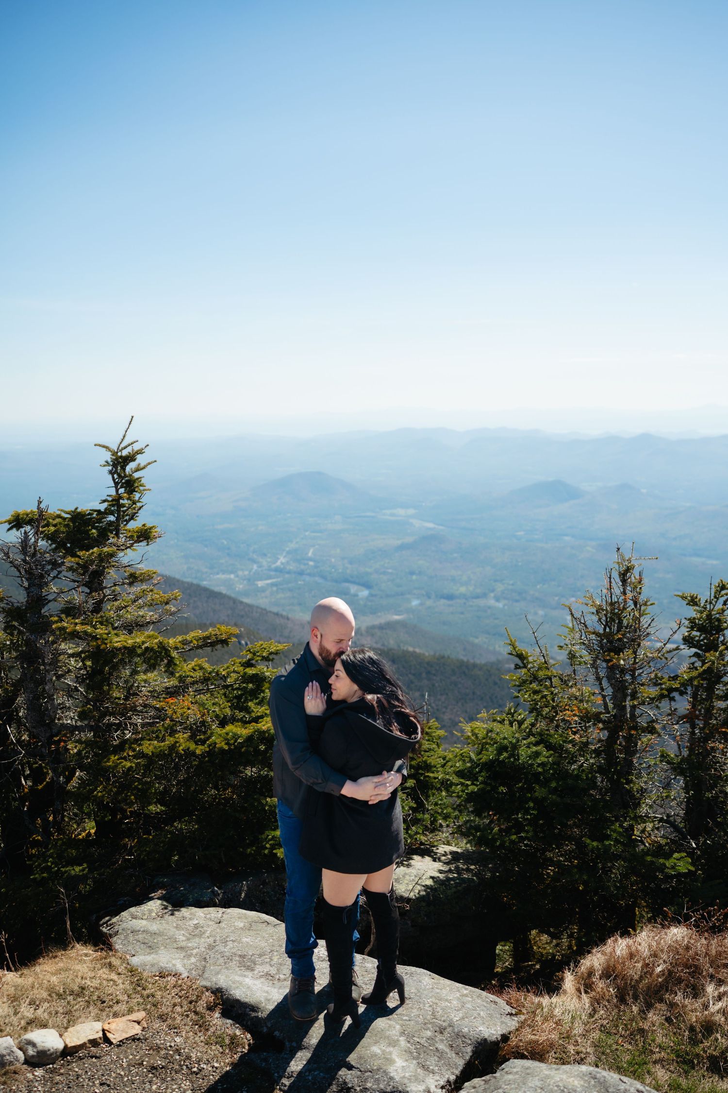 Couple embracing on rocky mountain summit with panoramic valley views and evergreen trees under clear blue sky.