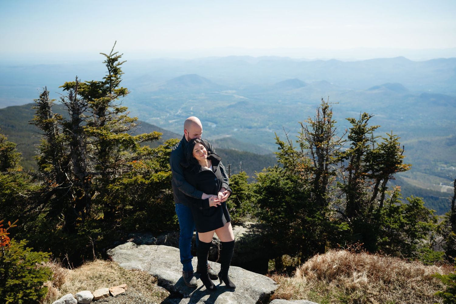 Couple standing together on mountain peak overlooking vast valley landscape with rolling hills and blue sky.