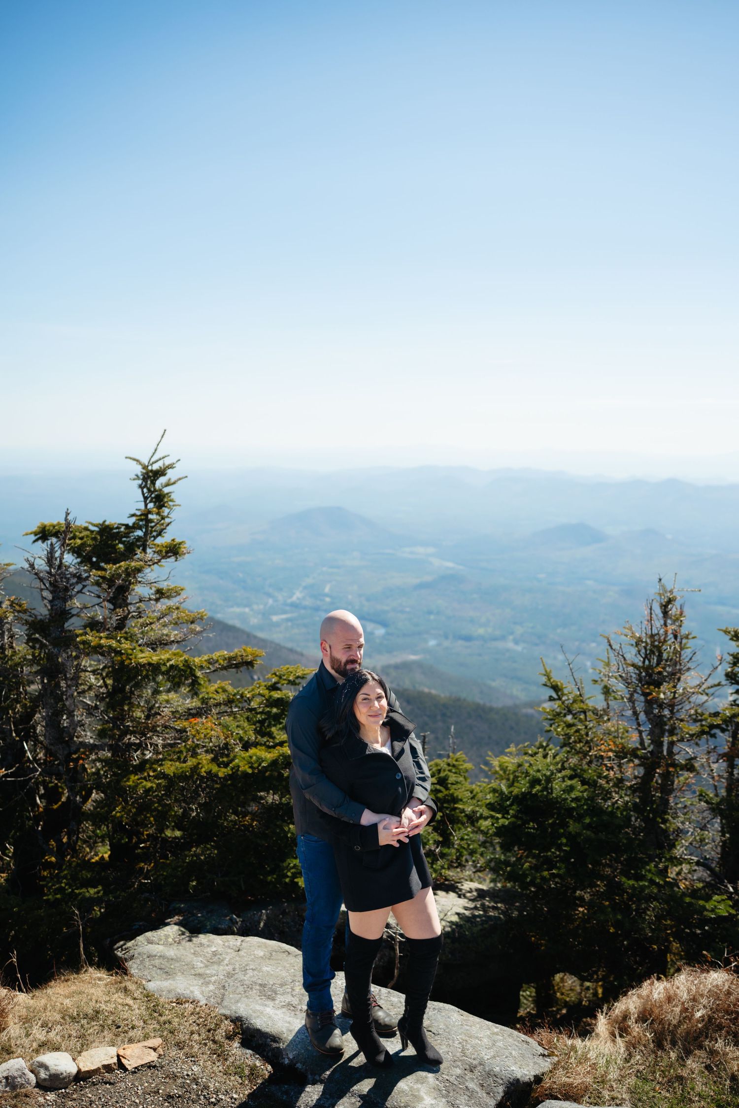 Couple embracing on a mountain summit with a vast scenic valley view behind them.