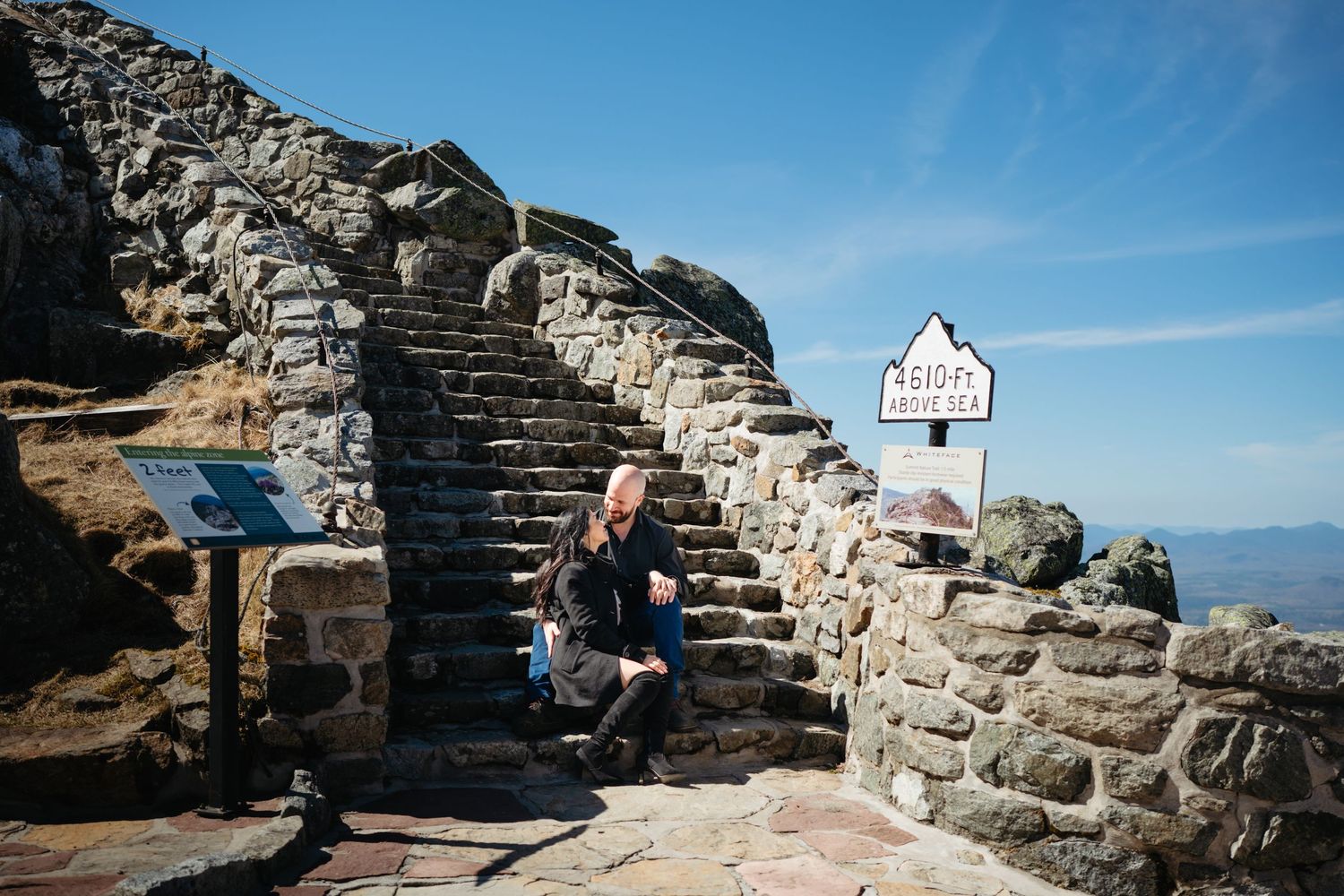 Person sitting on stone steps at a mountain summit marker under a clear blue sky.
