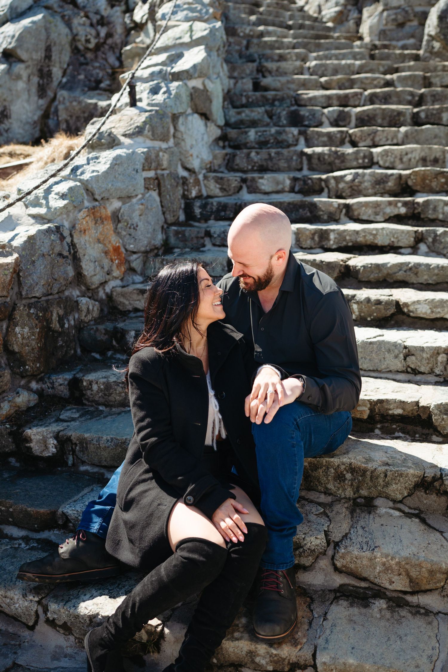 A couple sits close on ancient stone steps, smiling at each other, dressed in dark coats on a sunny day.