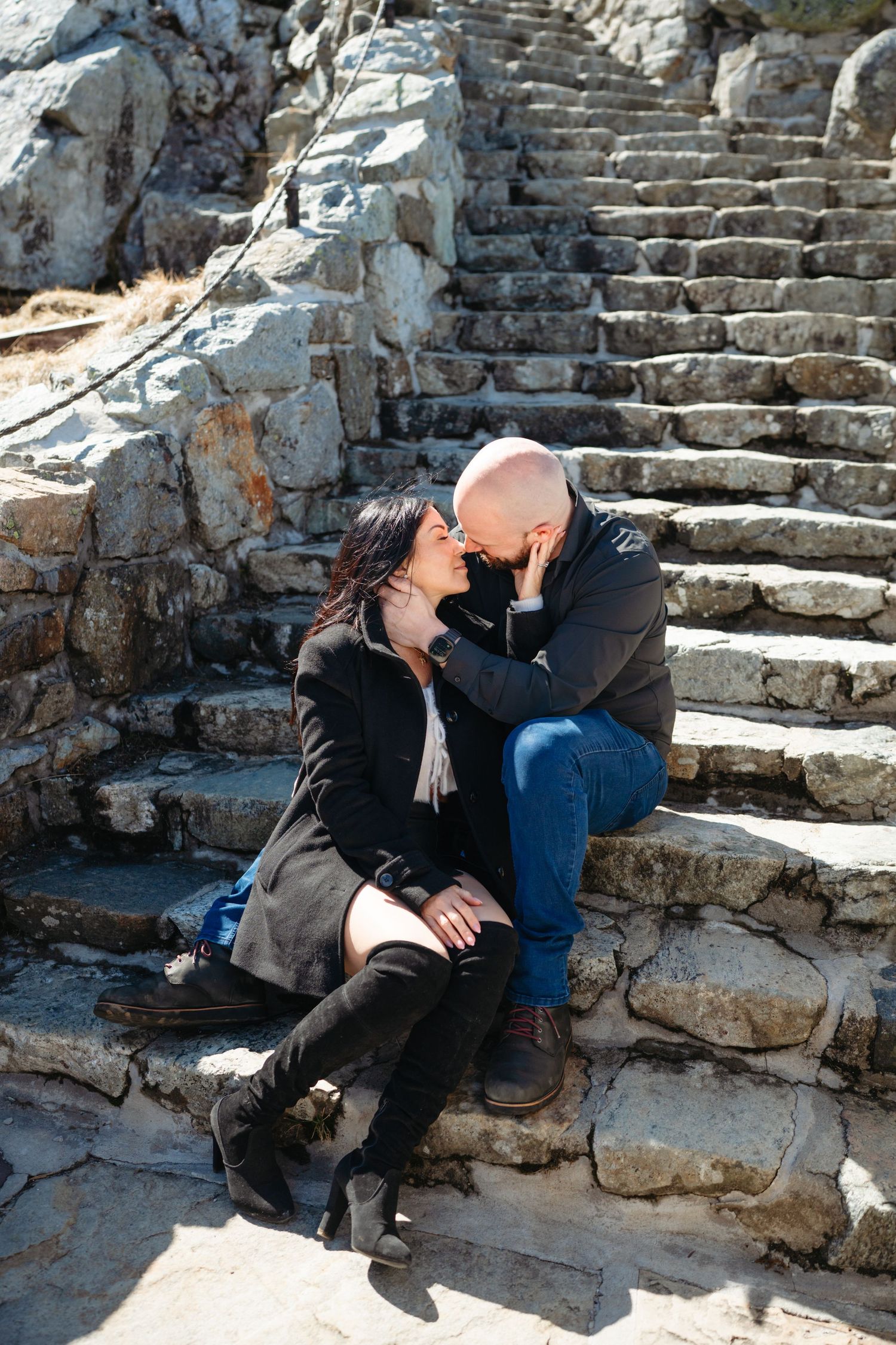 Couple sharing a romantic moment on ancient stone steps, woman in black coat and boots, man in jeans leaning in for a kiss.