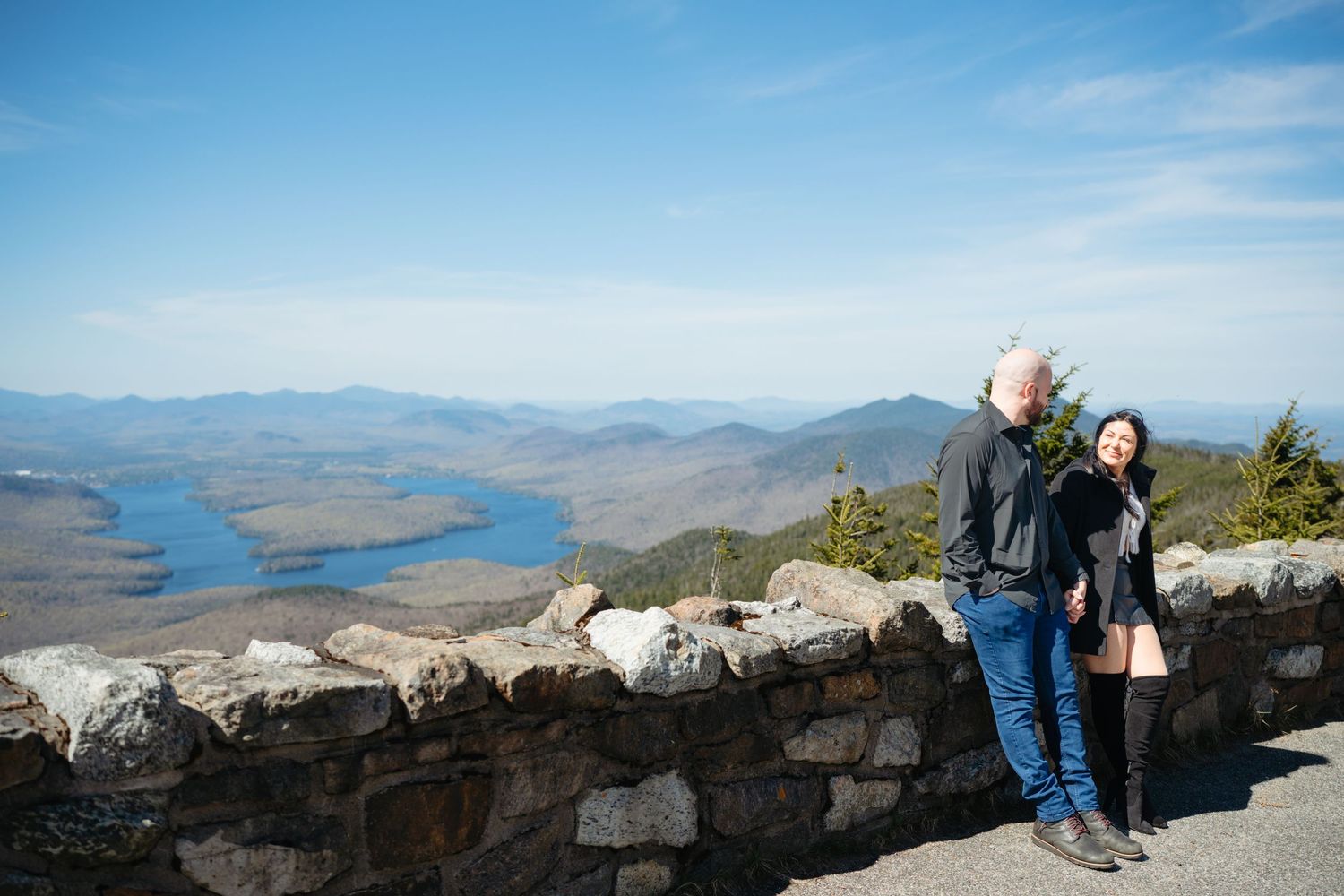 Couple standing at a stone overlook with stunning lake and mountain valley views on a clear day.