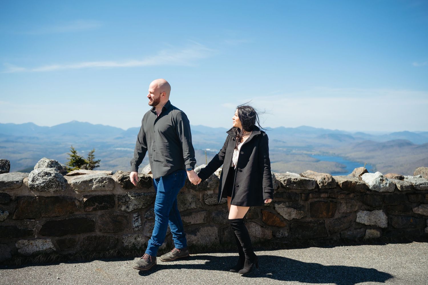 Couple walking hand in hand along a stone wall overlook with stunning mountain landscape and blue sky.