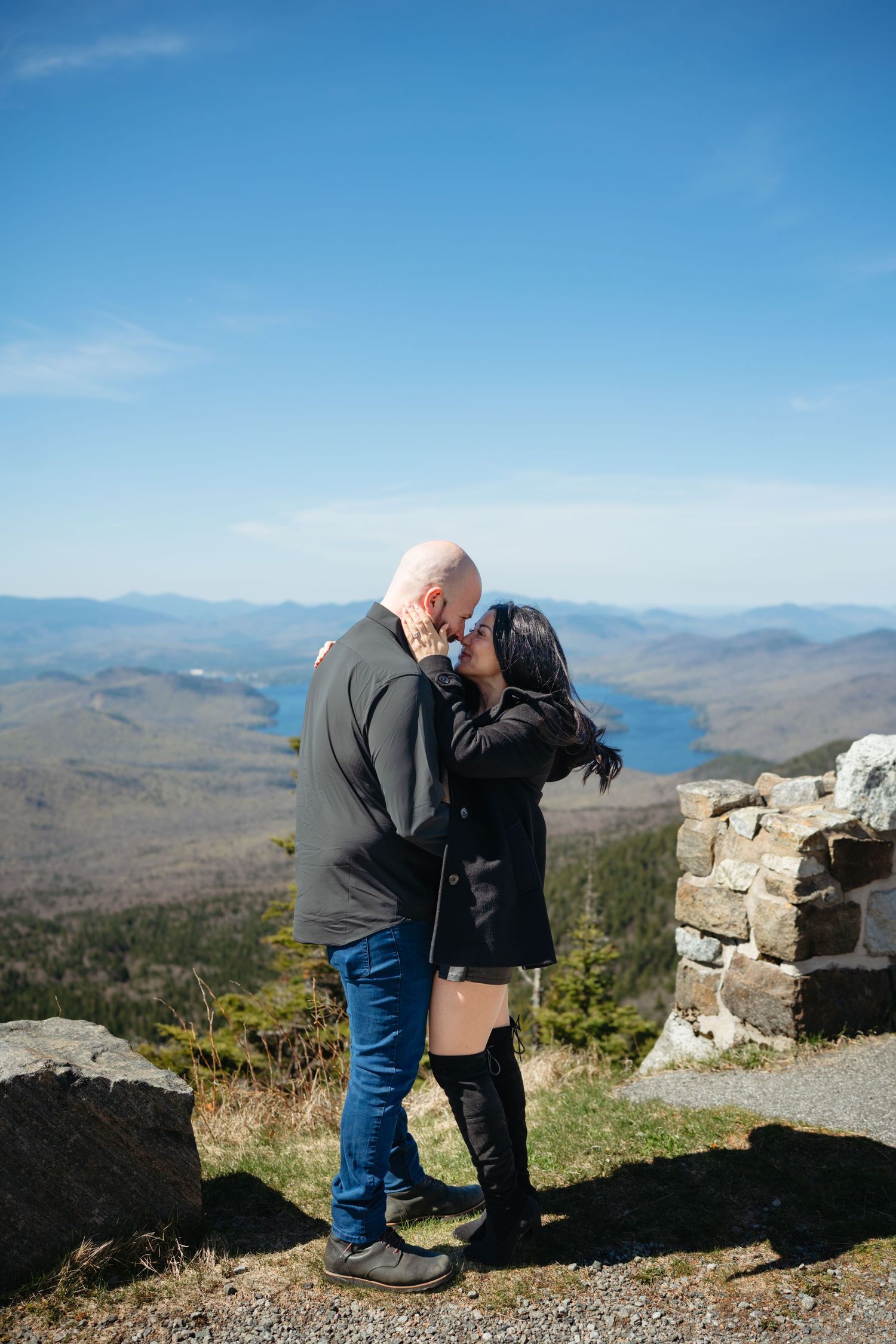 Couple sharing a kiss on a rocky mountain overlook with stunning lake and valley views behind them.