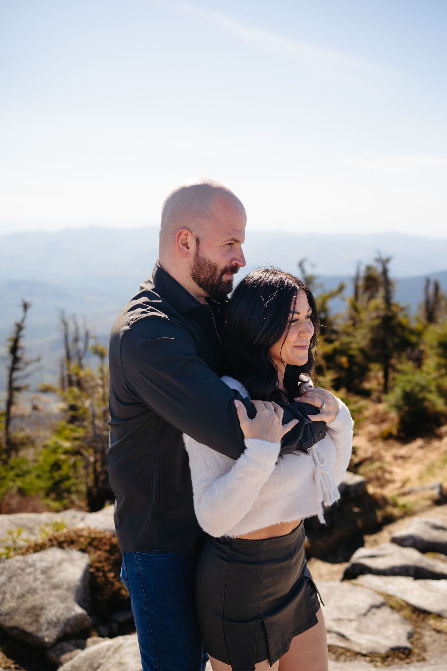 Couple embracing on a mountain summit with panoramic views, man in black jacket hugging woman in white sweater.