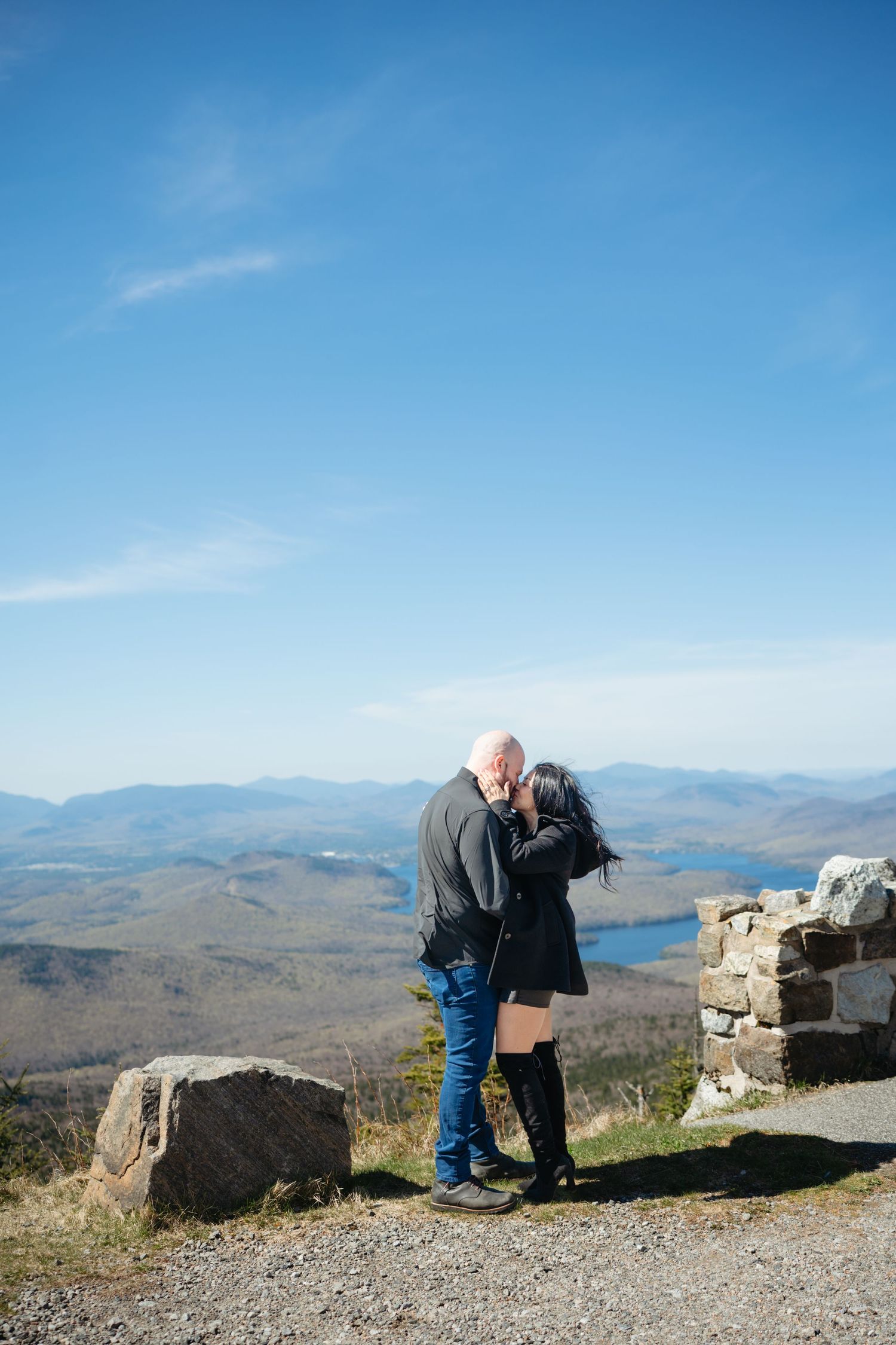 Couple sharing a romantic kiss on a mountain summit overlook with stunning blue sky and lake valley views.