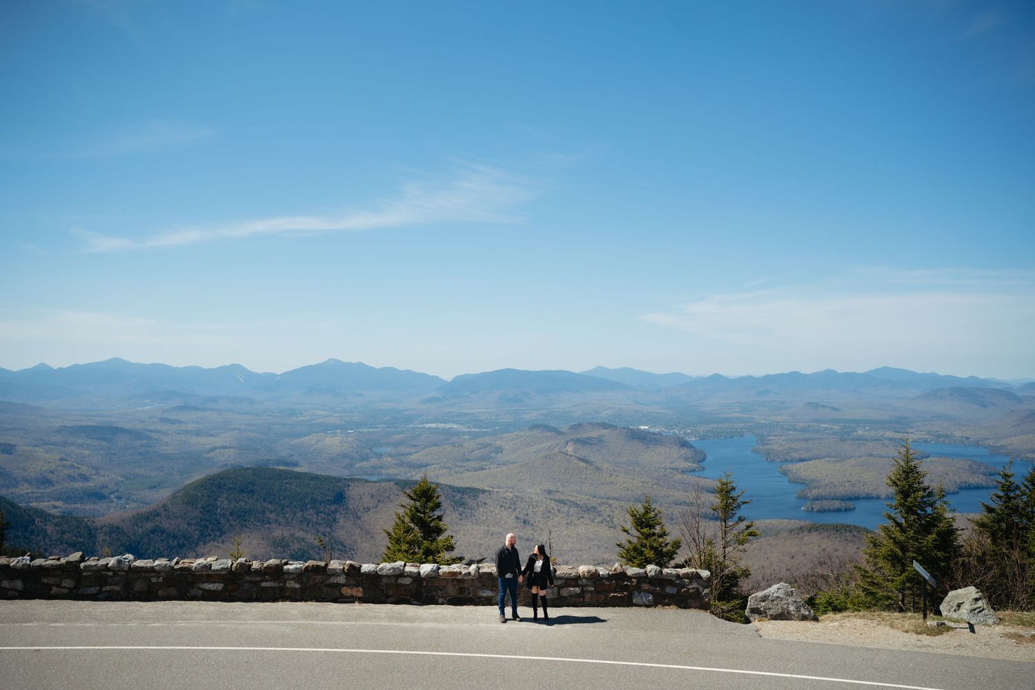 Two people stand at a mountain overlook with panoramic views of valleys, forests, and a lake under a clear blue sky.