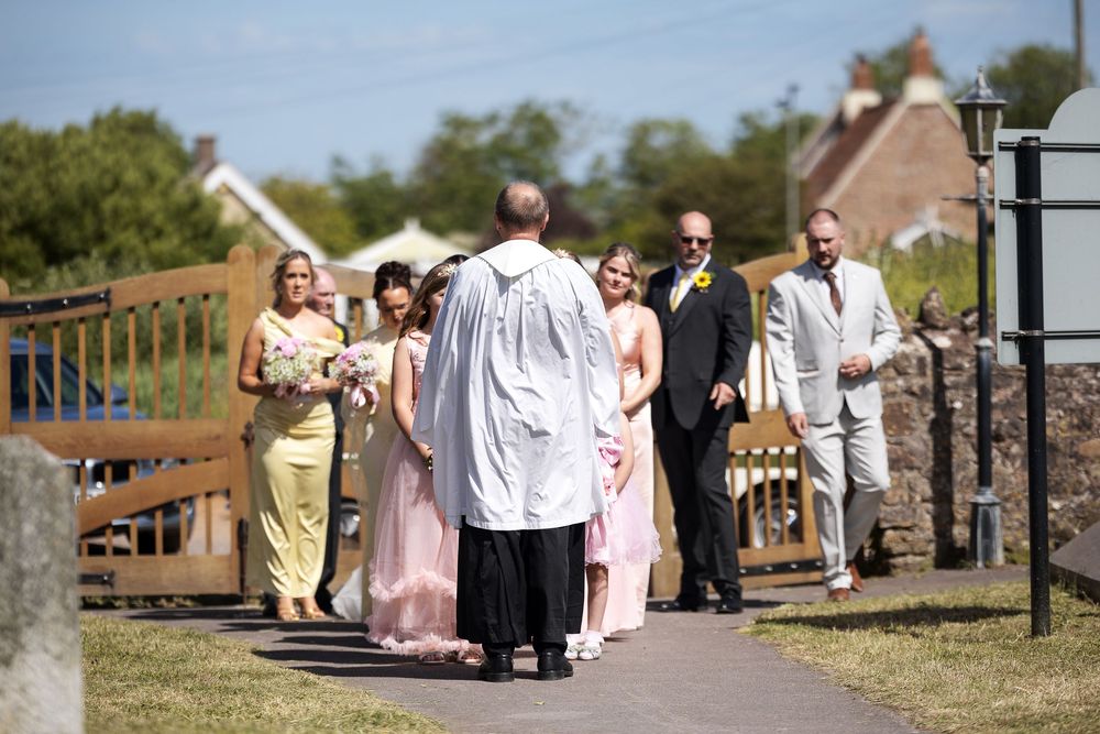 Wedding procession led by officiant in white robes, with bridesmaids in pink and yellow, walking along a church path.