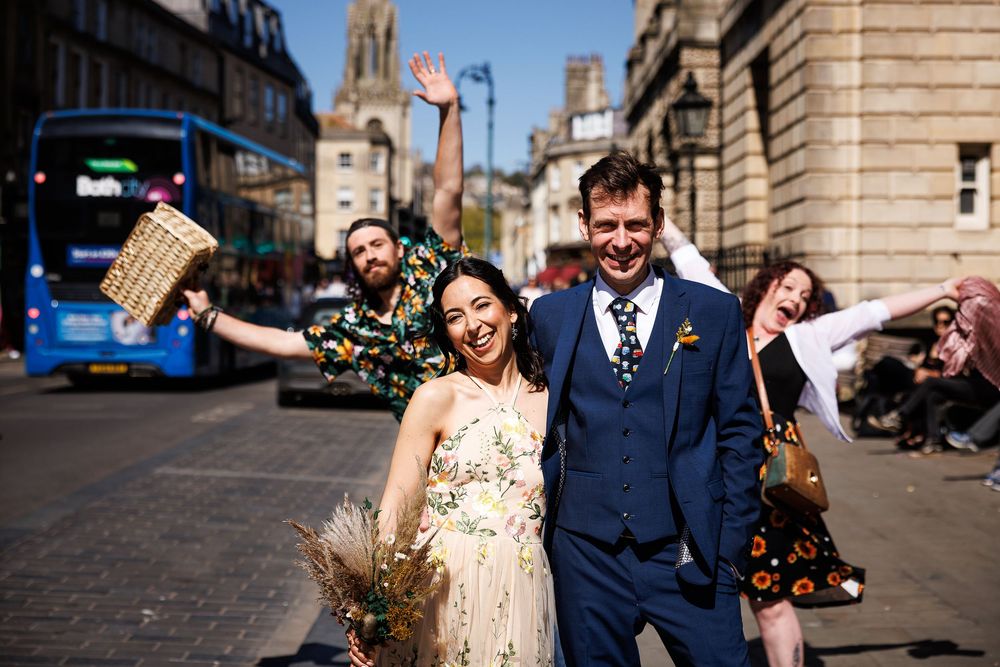 Joyful newlyweds celebrate on a sunny Bath street, bride in floral dress, groom in navy suit, friends cheering behind them.