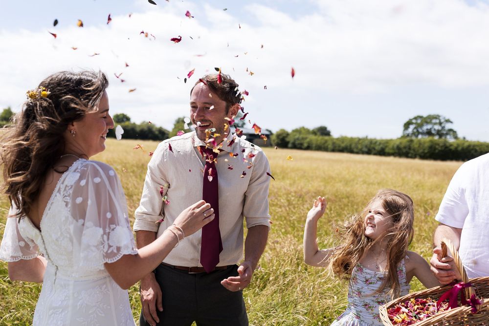 Bride and groom celebrating outdoors with confetti thrown by a young girl in a sunny field on their wedding day.
