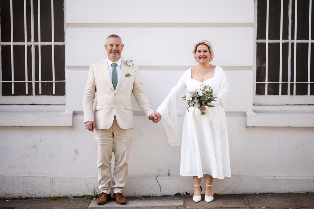 A smiling couple in wedding attire hold hands outdoors, the groom in beige suit and bride in white midi dress holding a bouquet.