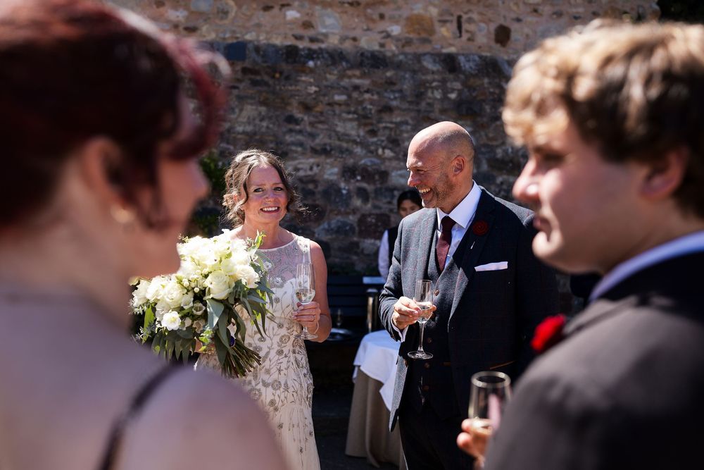 Smiling bride holding white bouquet chats with guests at outdoor wedding reception near stone wall.
