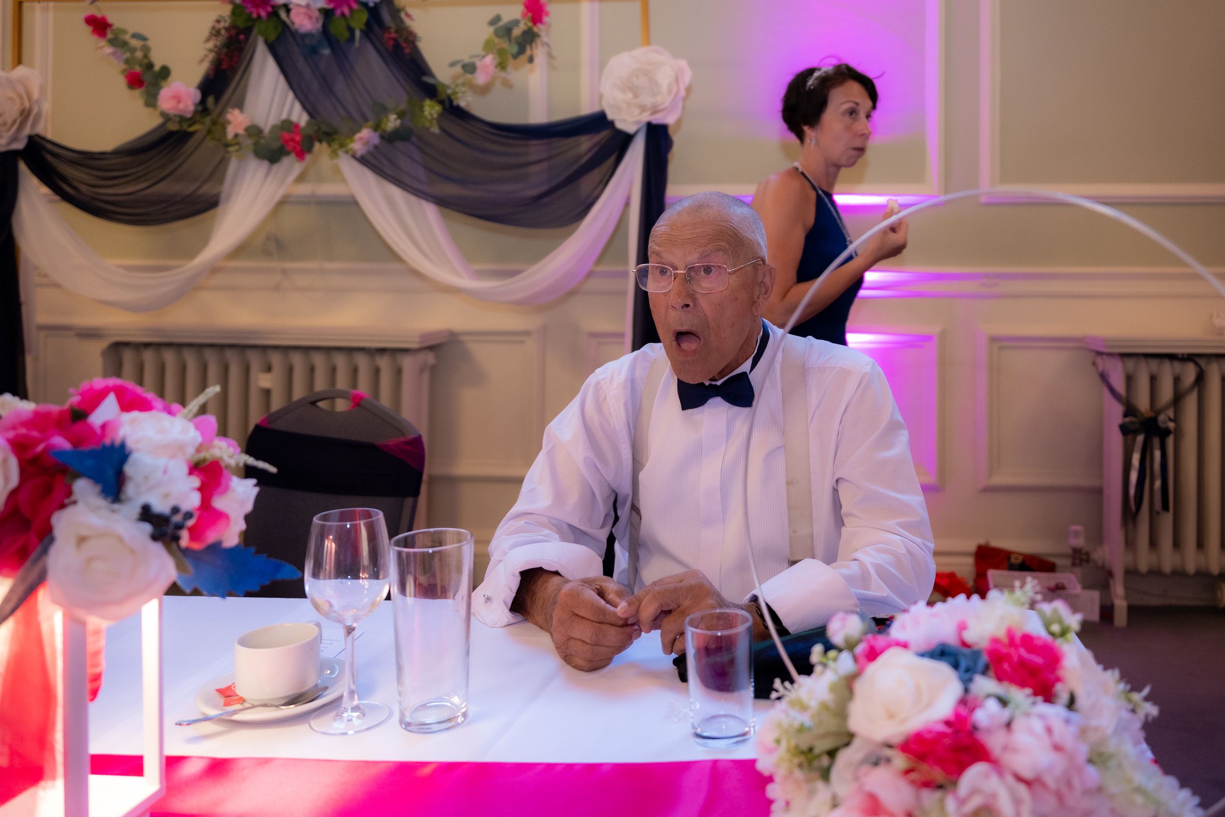 Elderly man in white shirt and bow tie seated at a decorated wedding reception table with colorful floral arrangements.