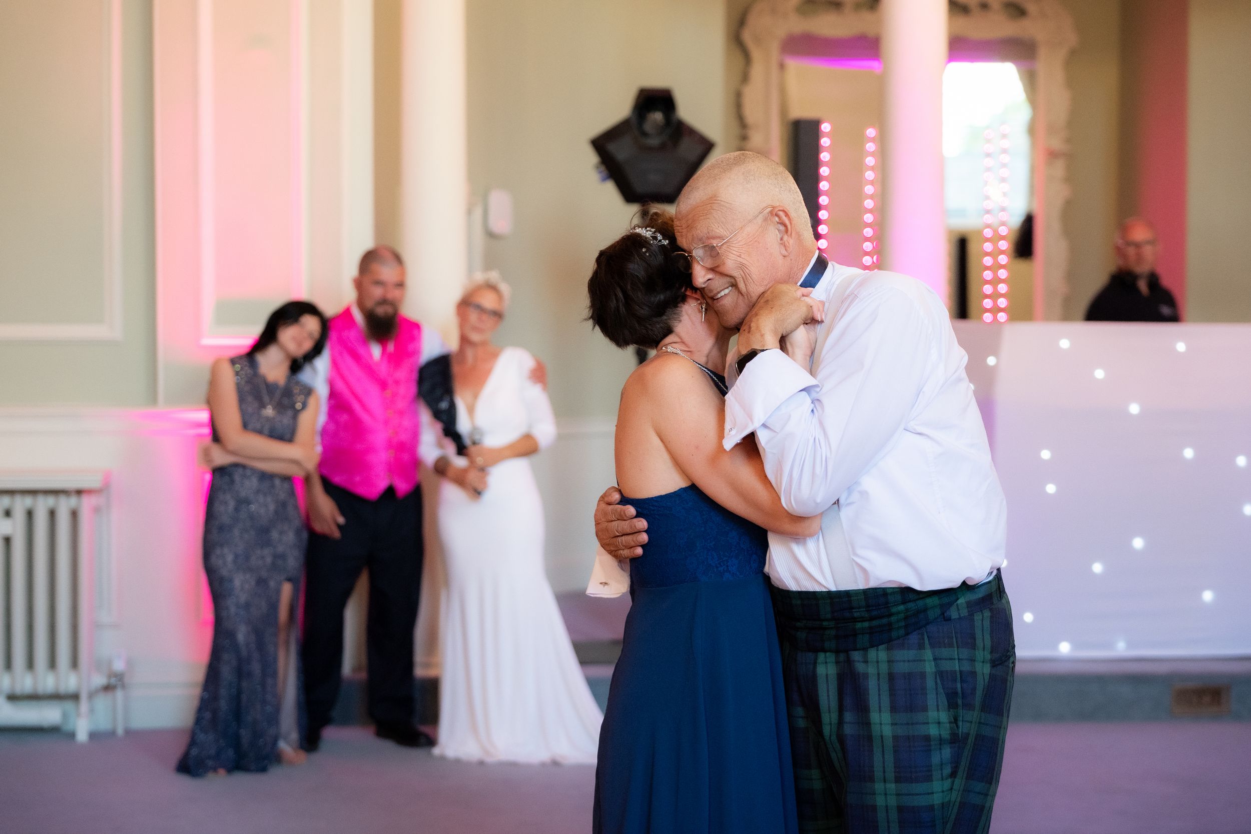 Father-daughter wedding dance at elegant venue with guests watching in background.