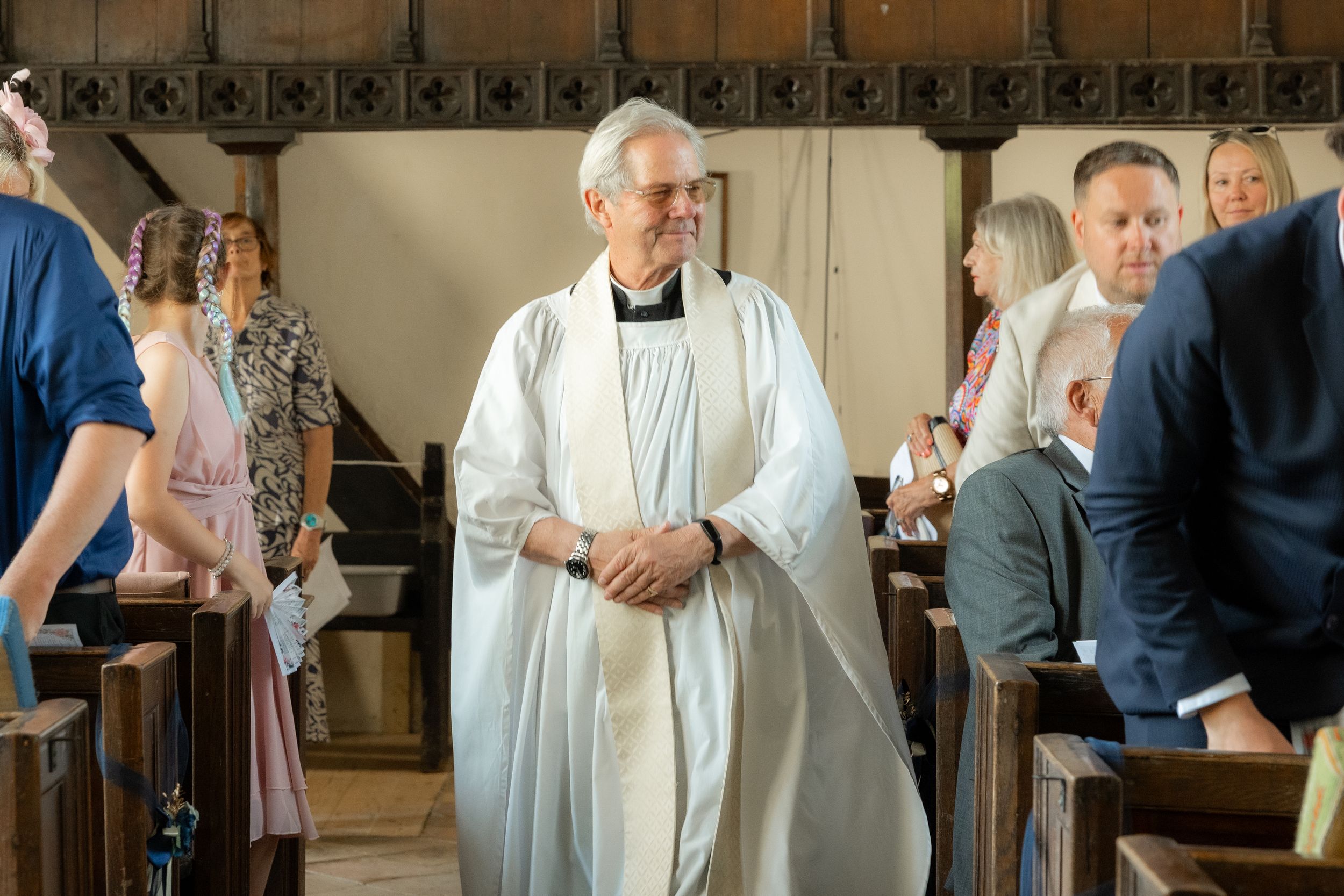 Smiling priest in white robes walks down the aisle of a traditional church during a wedding ceremony.