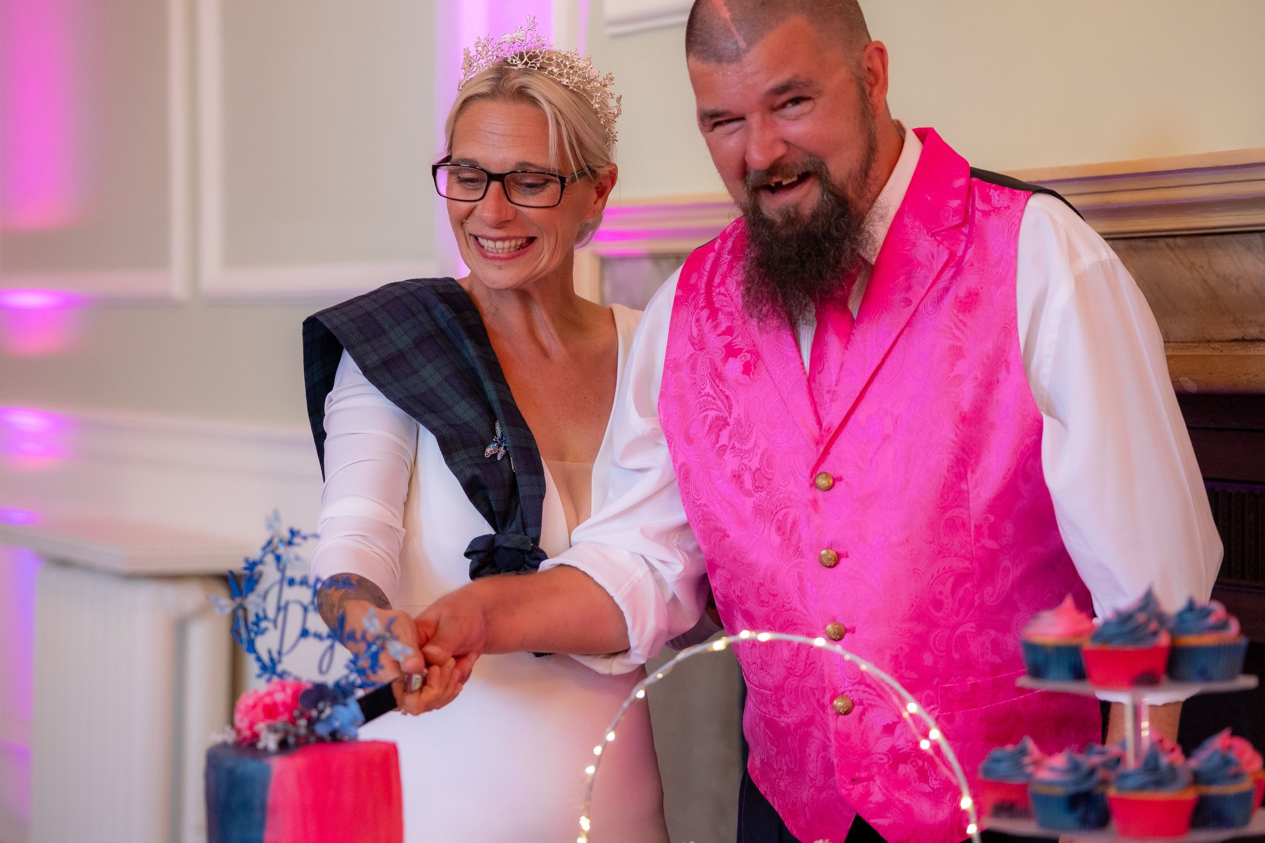 Bride and groom cutting their wedding cake, groom wearing a vibrant pink vest at a colorful reception.