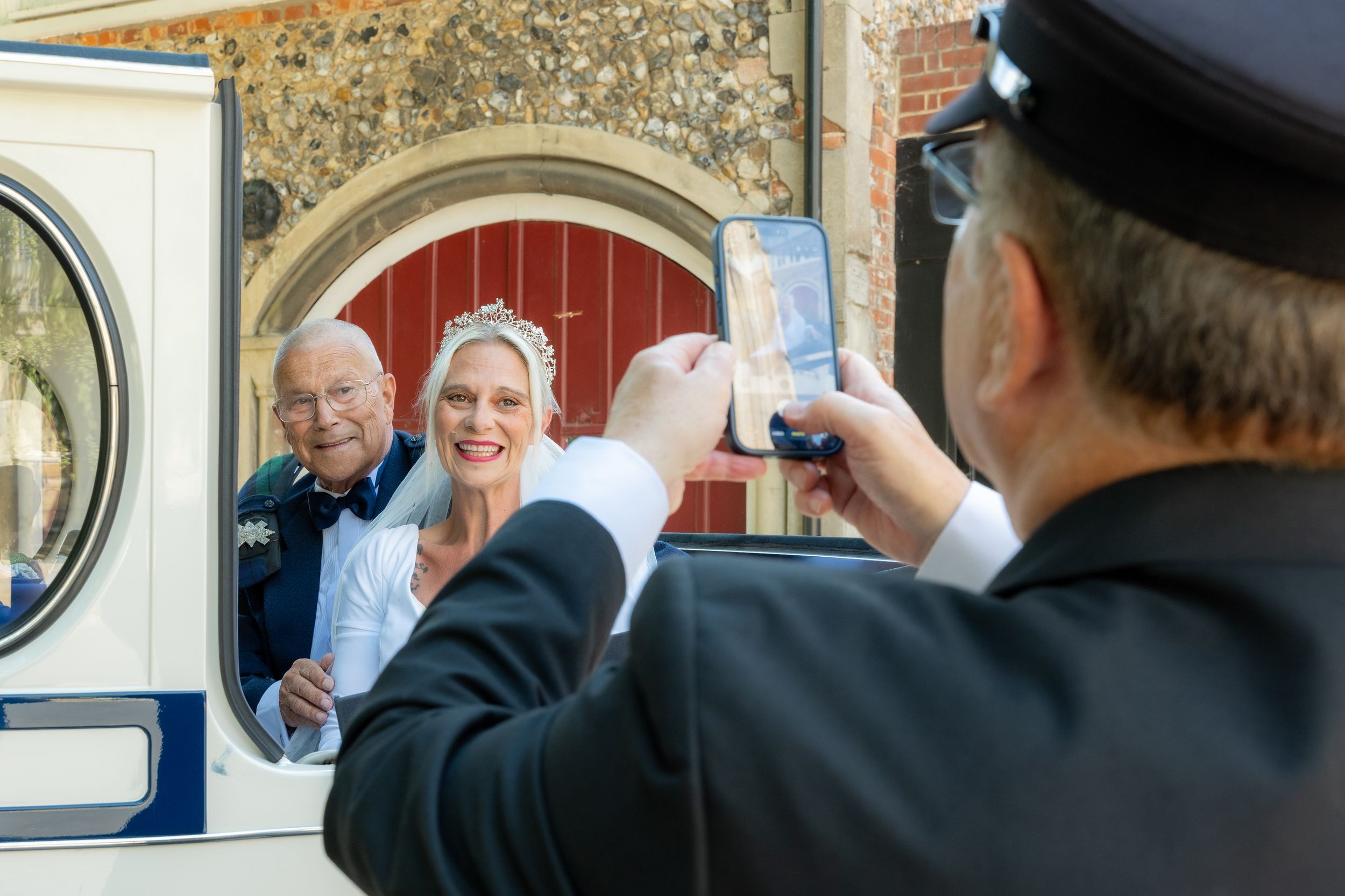 A chauffeur photographs a smiling bride and groom posing in a vintage wedding car doorway outside a church.