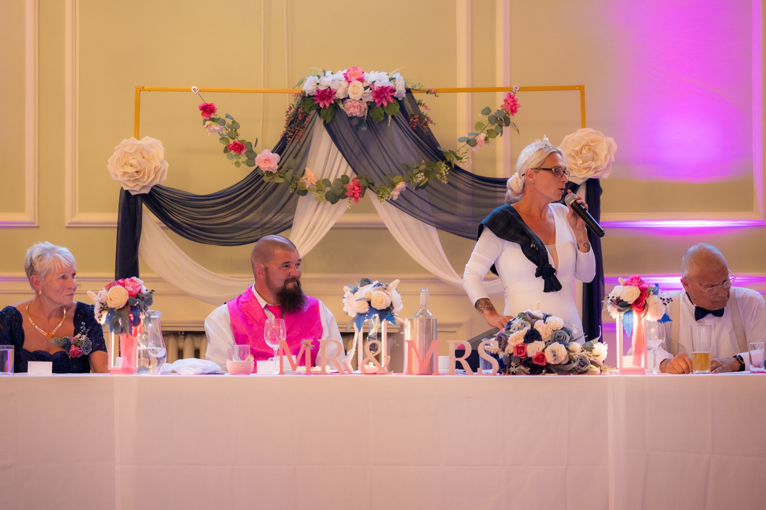 Woman gives a speech at an elegant wedding reception with floral decorations while guests sit at the head table.