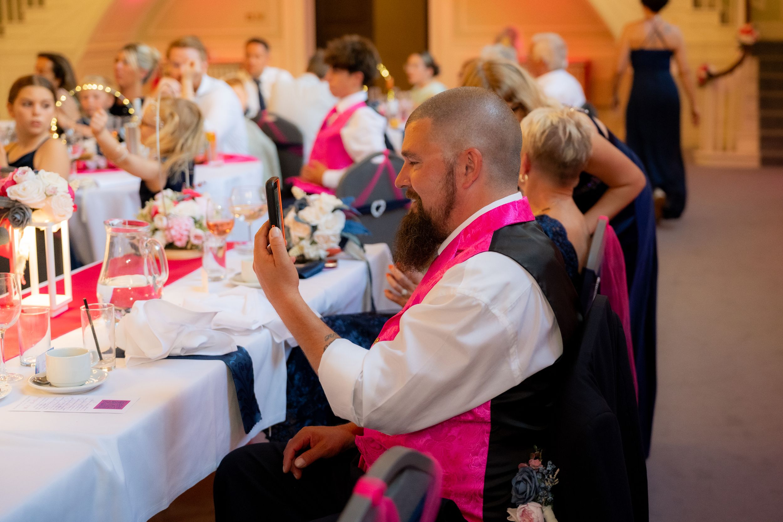 Bald bearded man in pink vest using smartphone at elegant wedding reception dinner with guests seated at decorated tables.