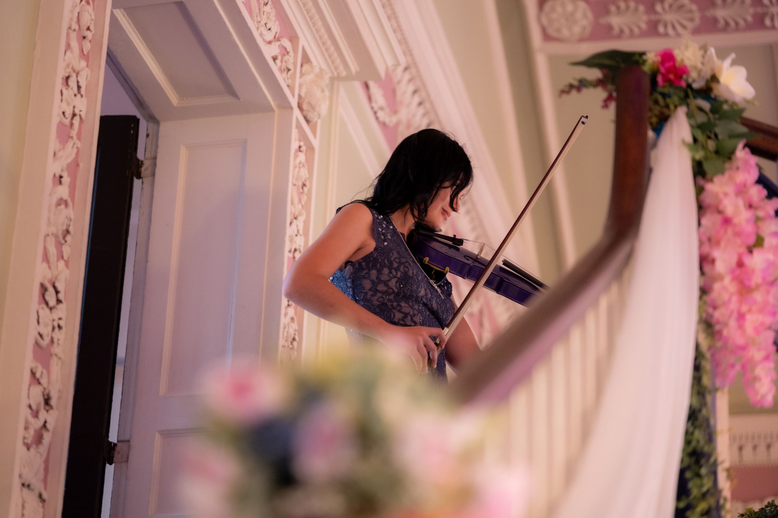 A woman in a blue dress plays violin beside a floral arch in an elegant decorated event venue.