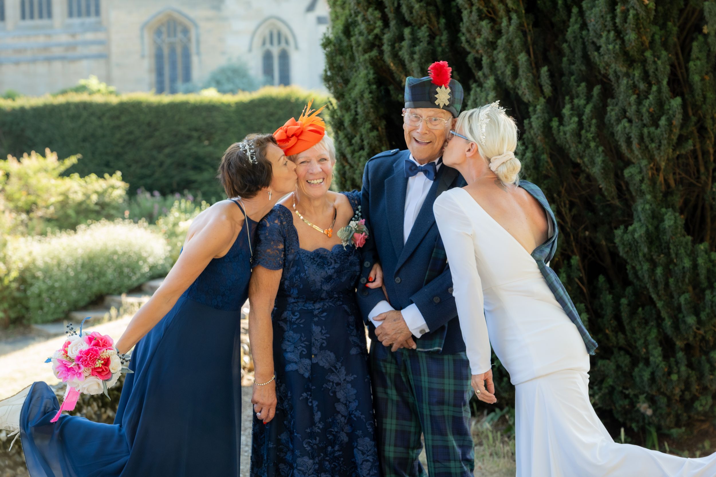 Wedding group photo with bride in white, groom in tartan kilt, and guests in navy blue outdoors.