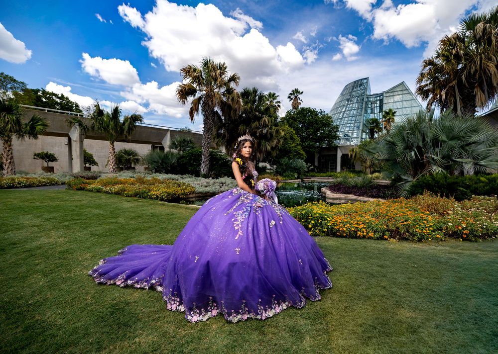 Teen in purple quinceañera ball gown posing in lush garden with palm trees and blue sky.