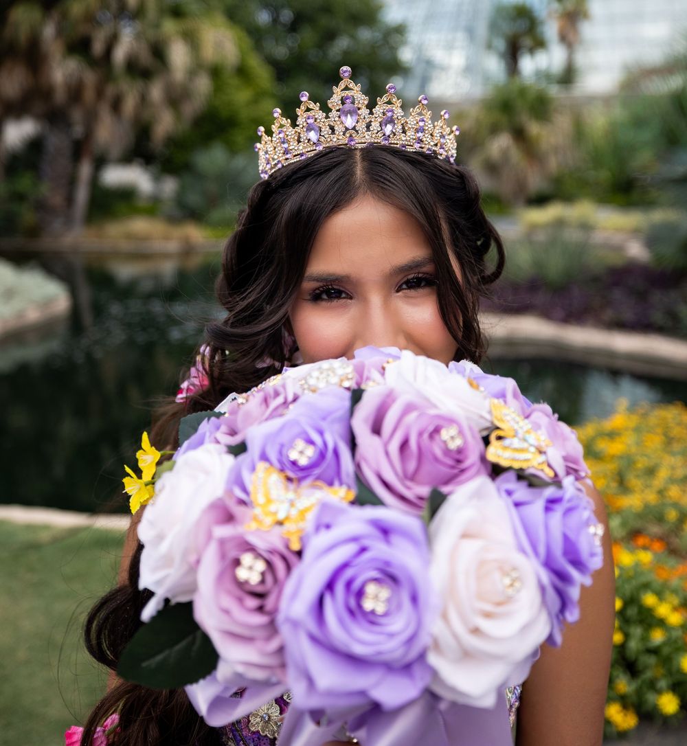 Young woman in a golden tiara holds purple and white roses bouquet at an outdoor garden celebration.