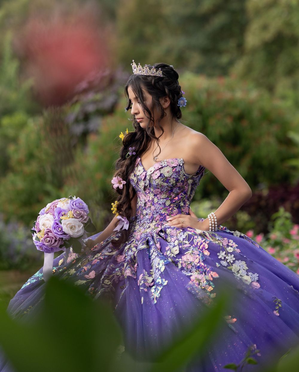 Young woman in purple floral quinceañera ball gown holding bouquet in colorful garden setting.