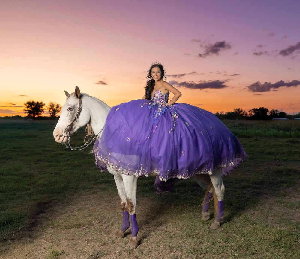 Young woman in purple quinceañera gown rides white horse at sunset in open field.