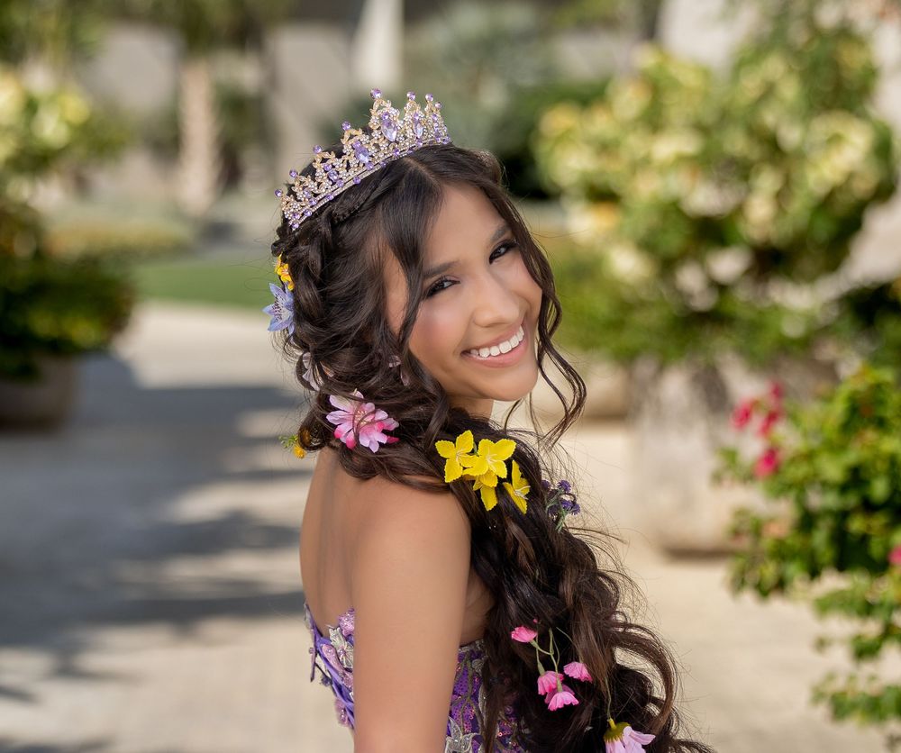 Smiling young woman wearing a purple gown, floral hair accessories, and a tiara at an outdoor quinceañera celebration.
