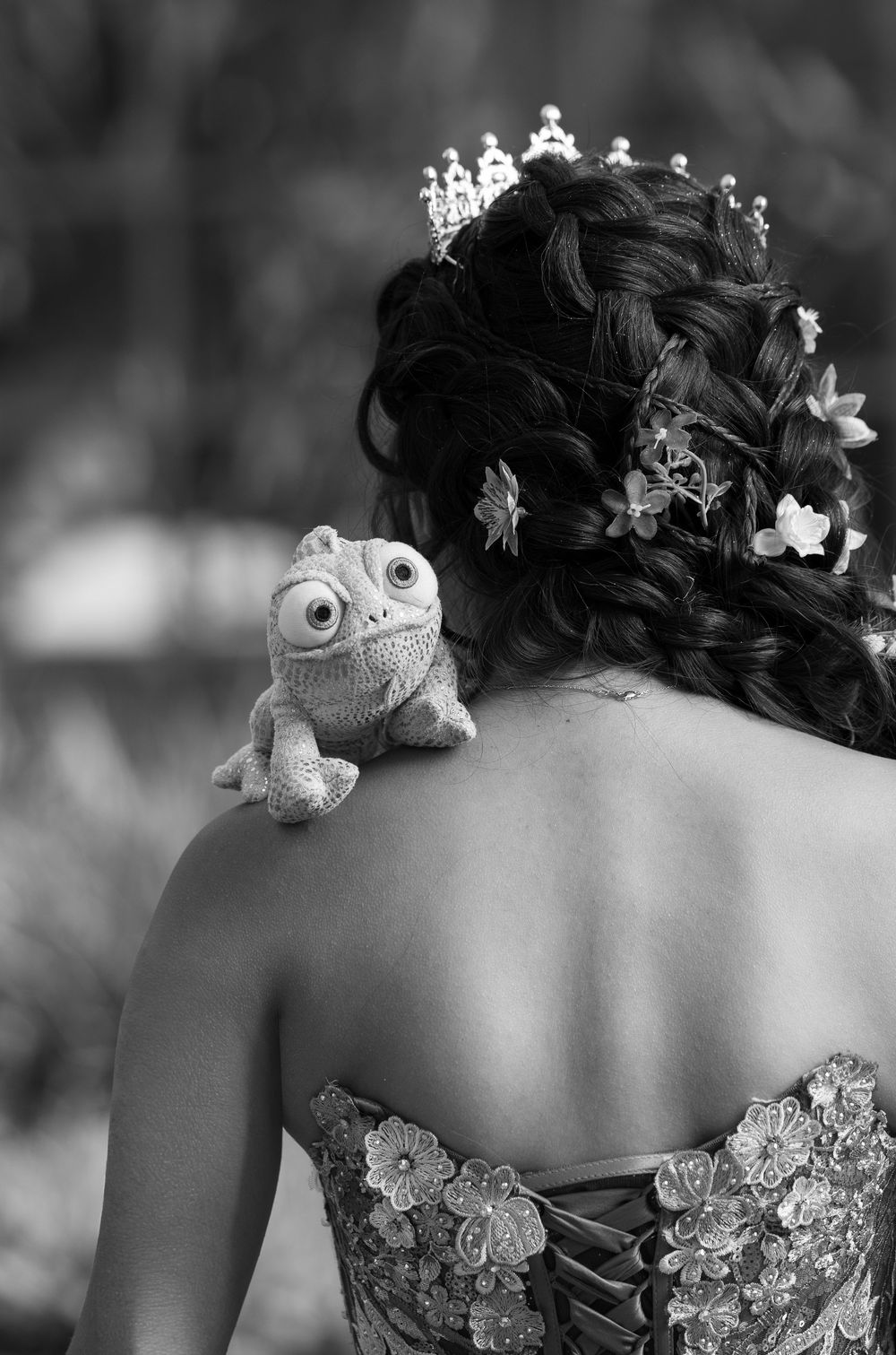 Black and white photo of quinceañera girl with braided updo, tiara, flowers, and Pascal chameleon plush on shoulder.