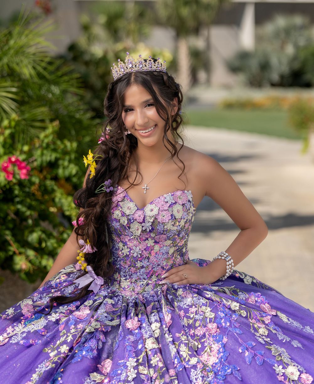 Young woman in purple floral quinceañera ballgown and tiara smiling outdoors.