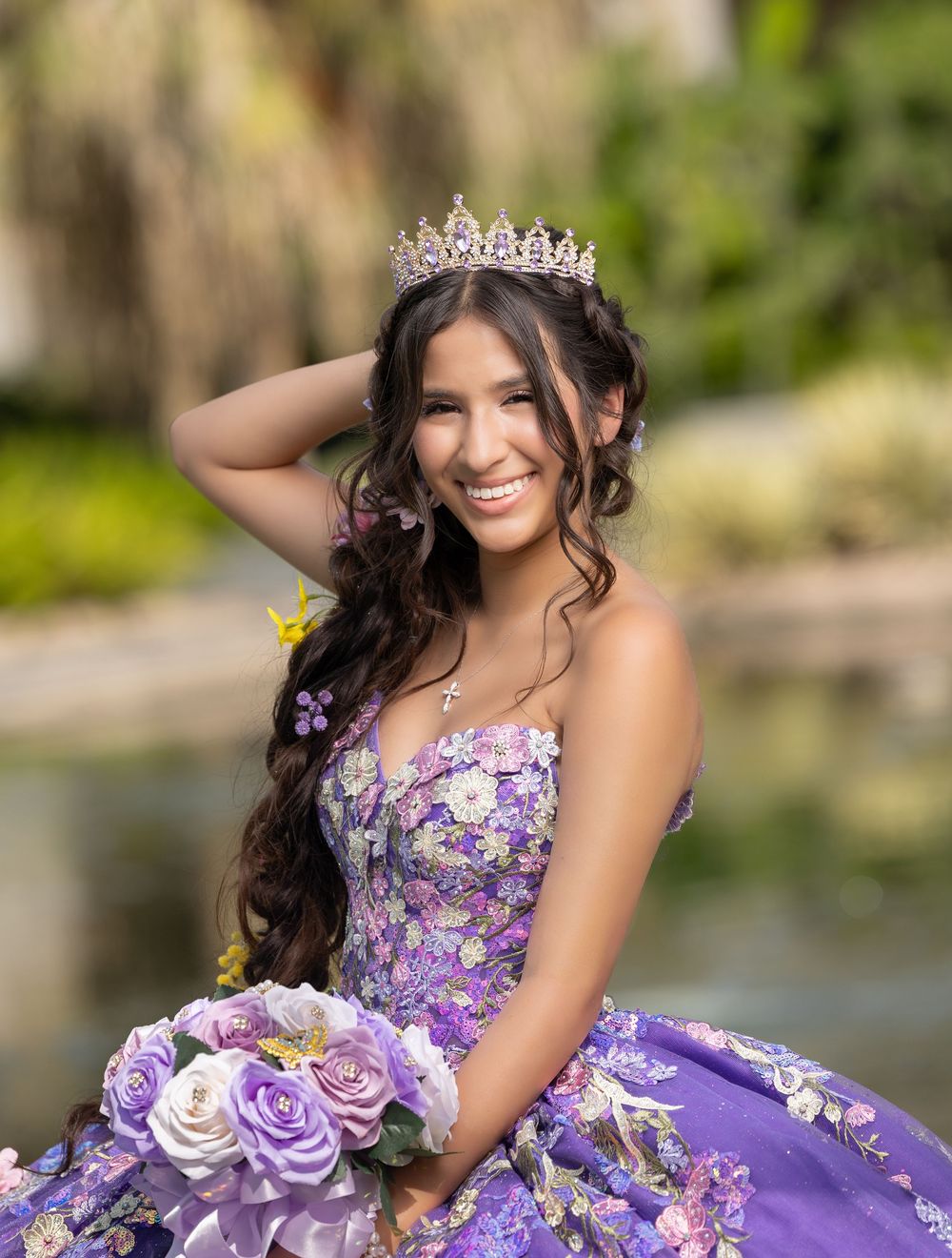 Young woman in purple floral quinceañera gown with tiara and bouquet smiling outdoors.