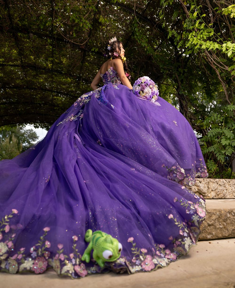Girl in purple floral quinceañera ball gown with long train posed under green trees outdoors.