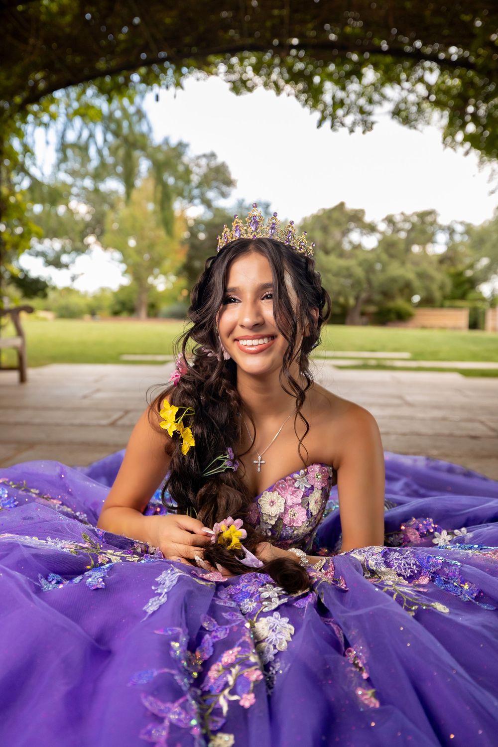 Smiling young woman in purple quinceañera gown wearing tiara holding yellow flowers in park.