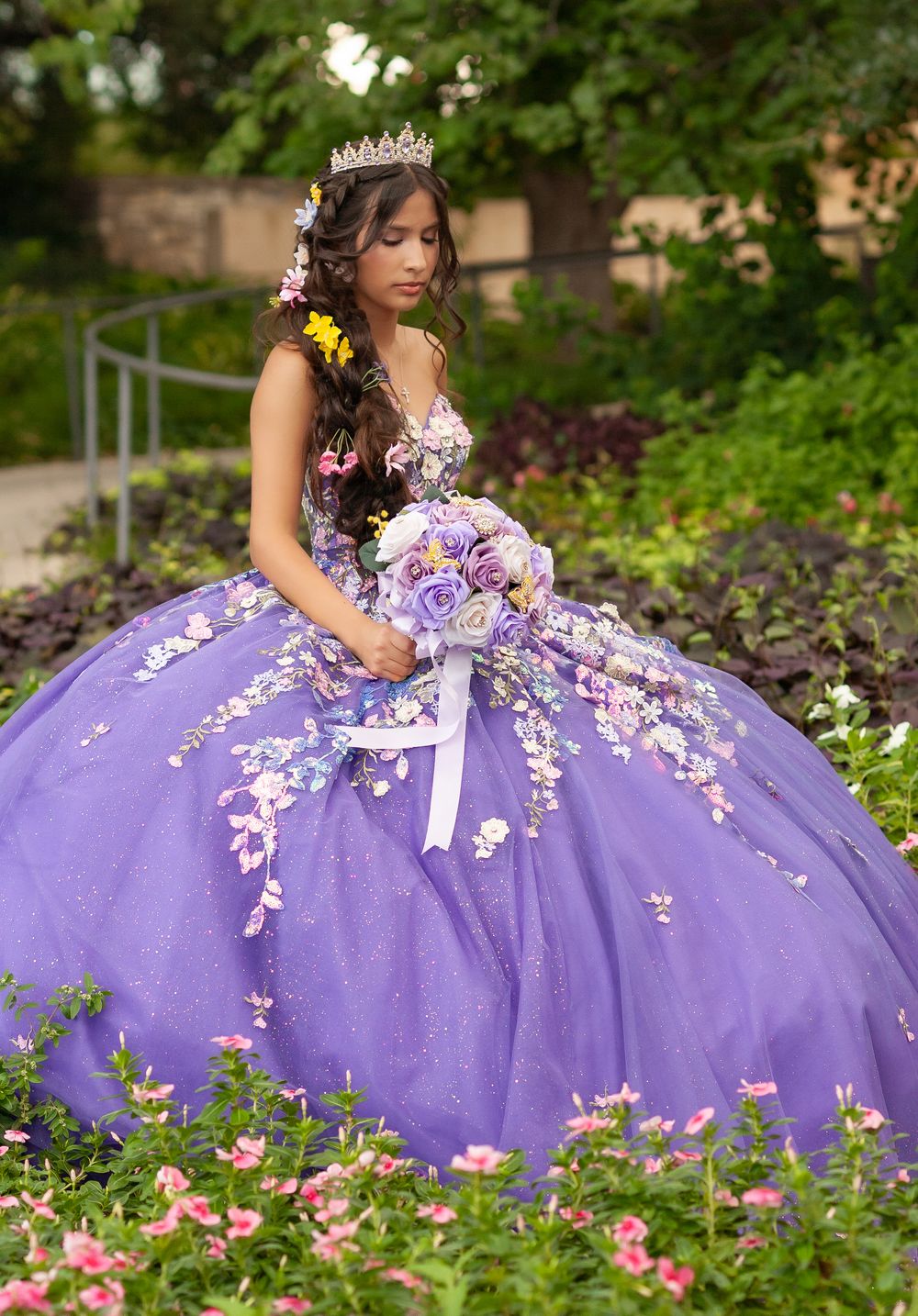 Young woman in purple quinceañera ball gown with floral appliqués holds bouquet in garden.