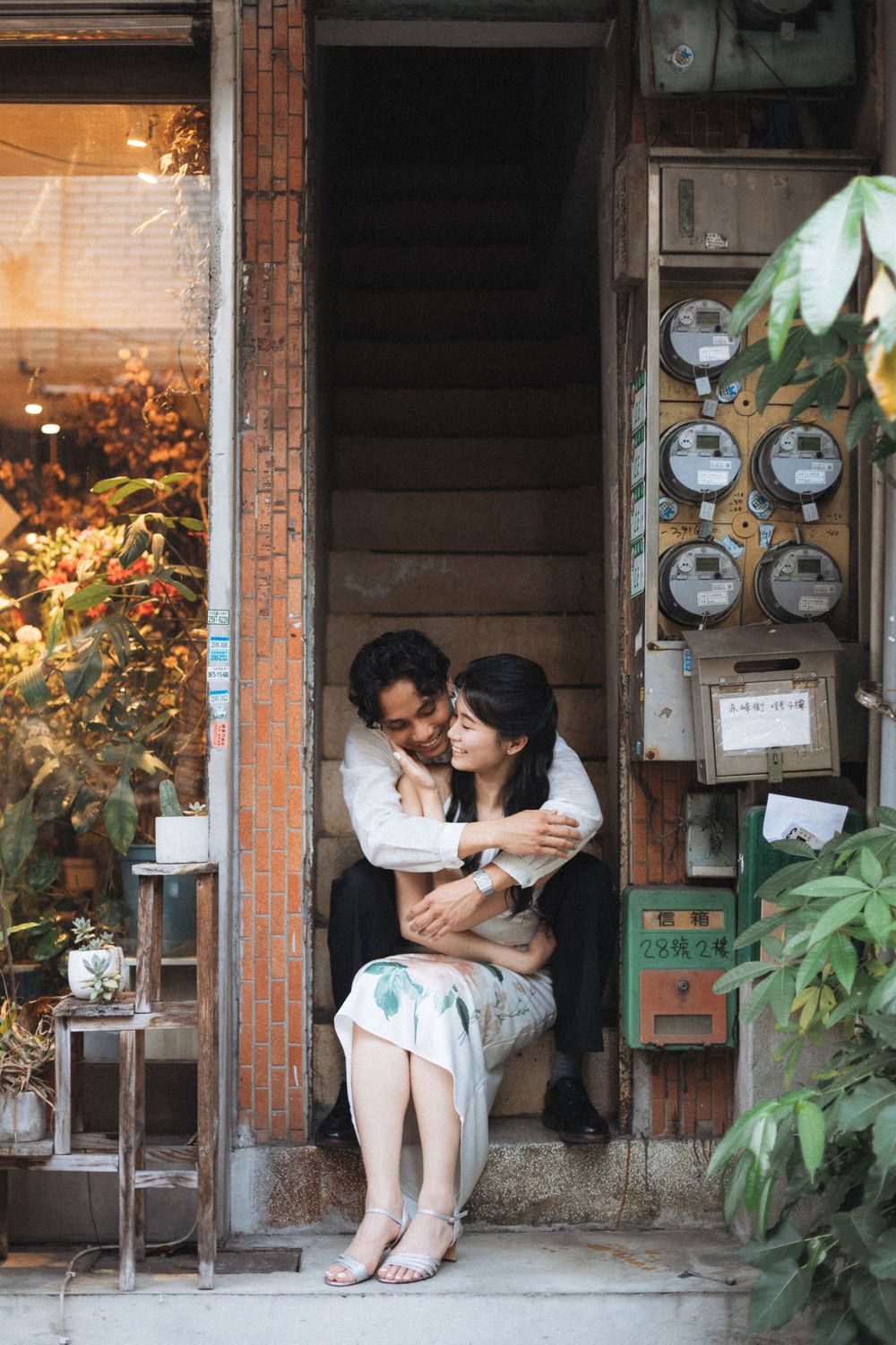 Couple sitting together in a vintage doorway, sharing a tender embrace near electric meters and lush greenery.