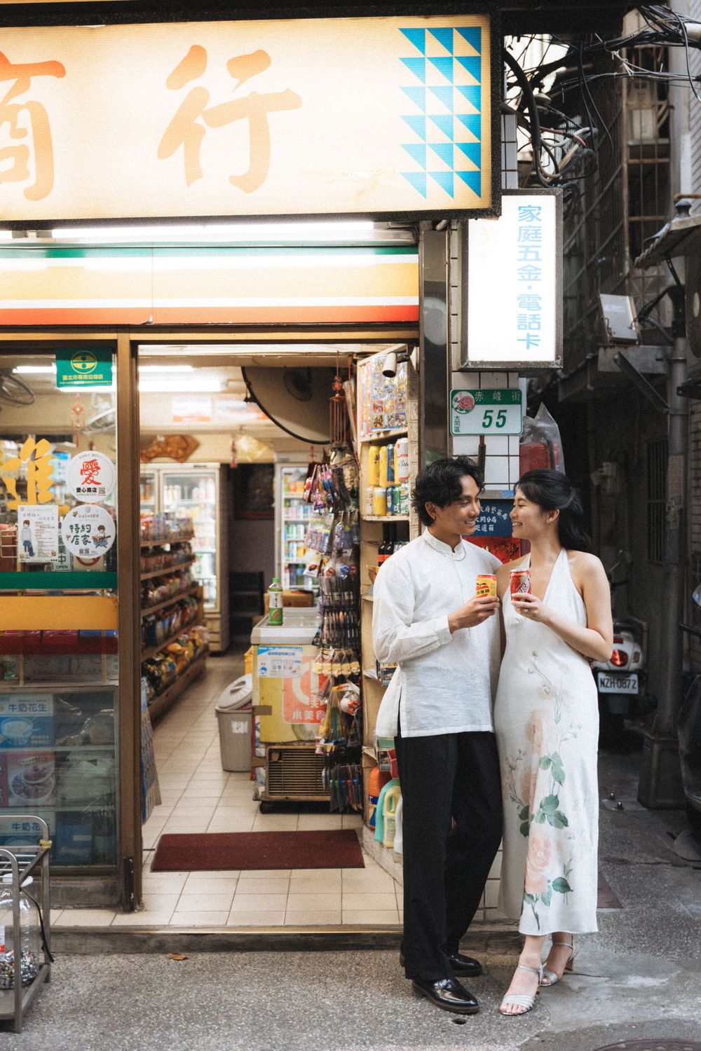 Couple sharing drinks outside a traditional Asian convenience store with Chinese signage at number 55.