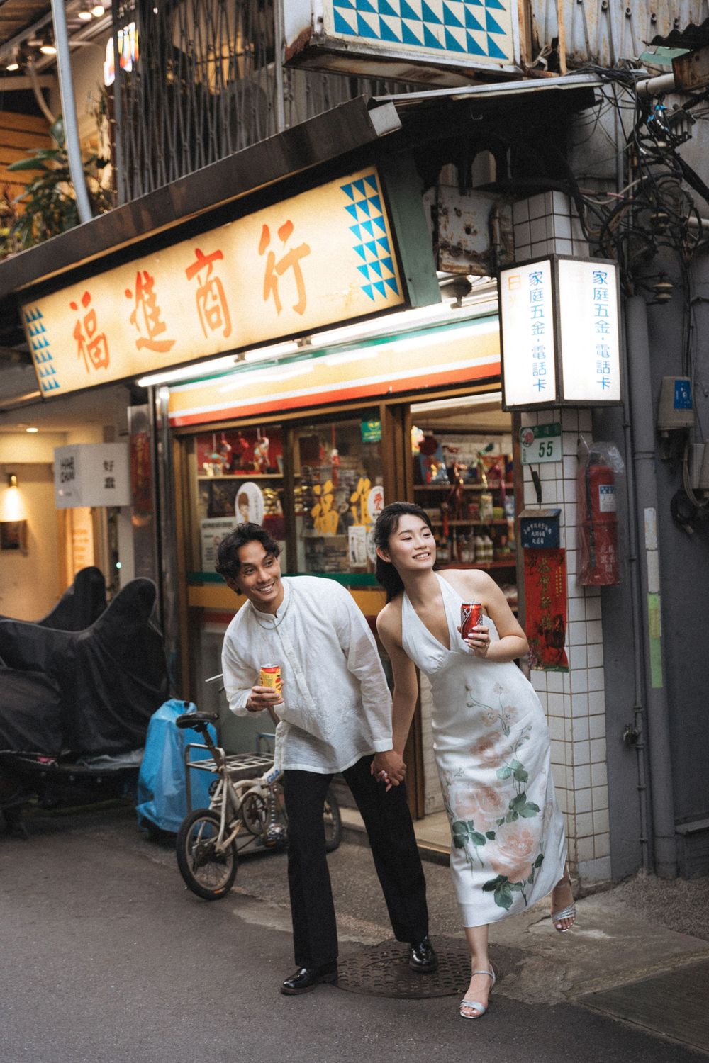 Couple holding drinks and smiling outside a traditional Chinese shop in an urban alley, dressed in elegant attire.