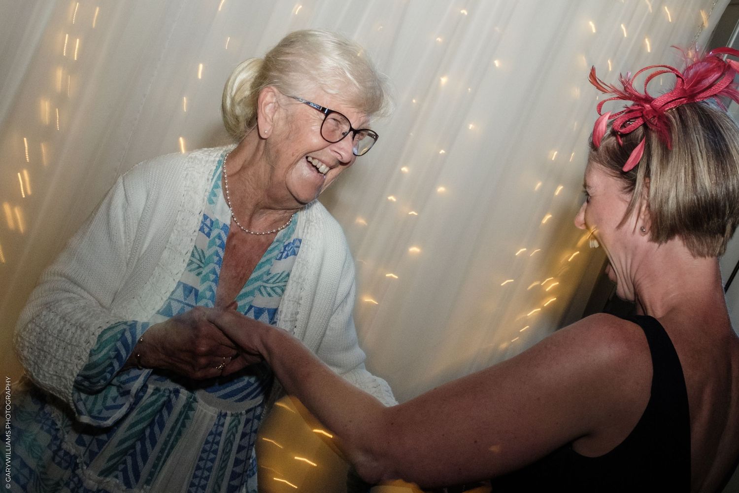Two women laughing and holding hands at a festive event with twinkling fairy lights backdrop.