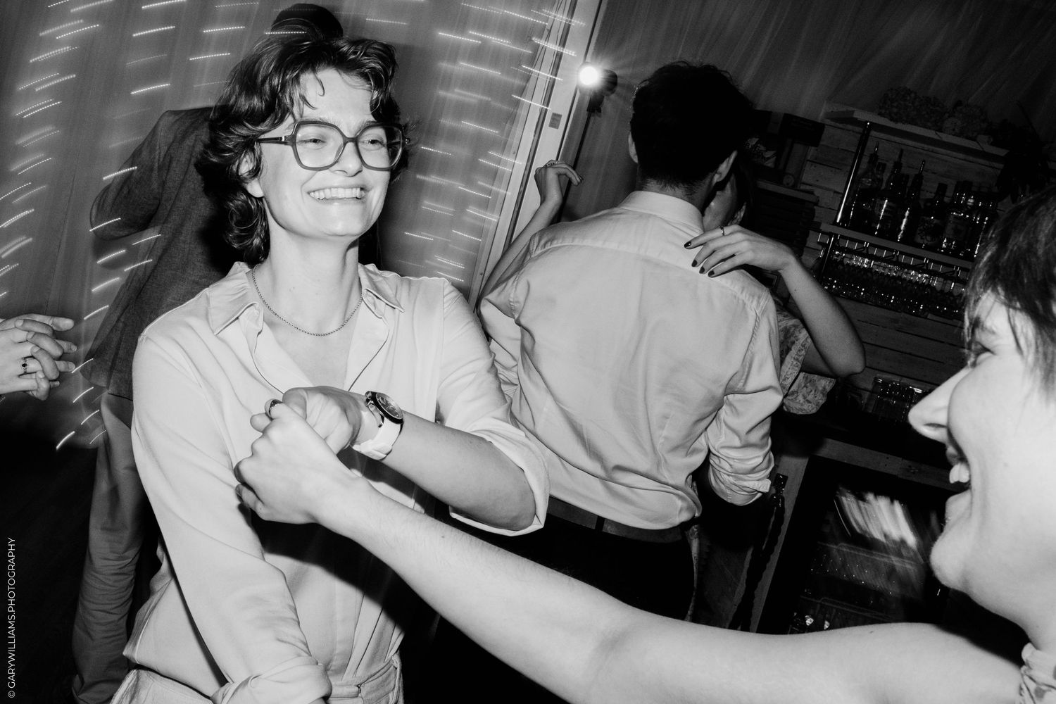 Black and white photo of a smiling woman with glasses dancing at a party, holding hands with another guest.