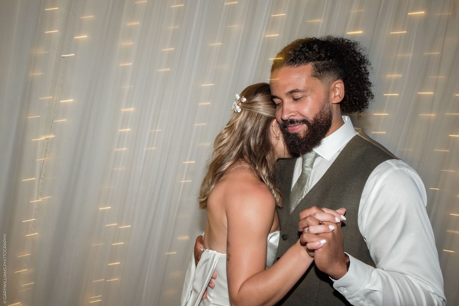 Bride and groom share first dance, holding hands closely in front of a twinkling fairy light backdrop.